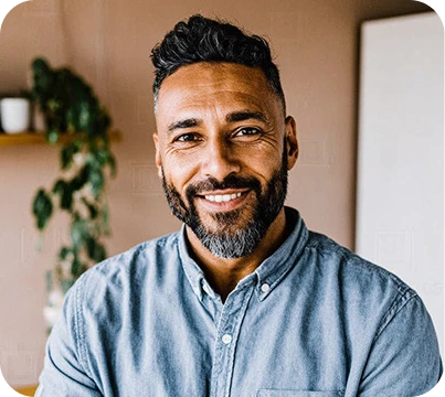 Smiling man with short curly hair and beard wearing a blue button-up shirt indoors with plant in background.