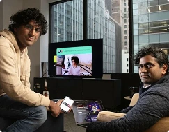 Two men sitting in a modern office setting with cityscape windows, one holding a smartphone and the other using a laptop showing a video call with a woman.