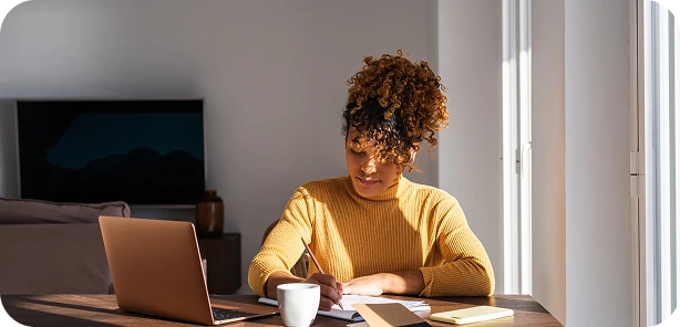Woman with curly hair in a yellow sweater writing at a table with a laptop, coffee cup, and notebook near a window.