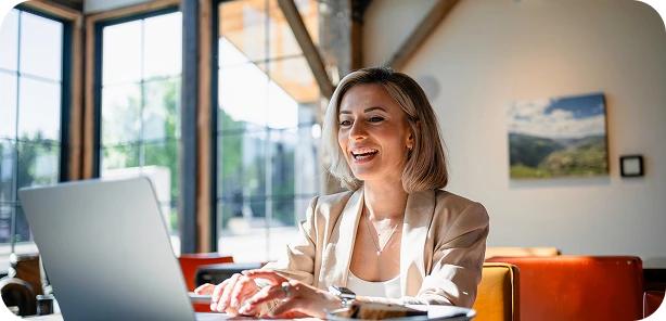 Smiling woman working on a laptop in a bright, modern cafe with large windows.