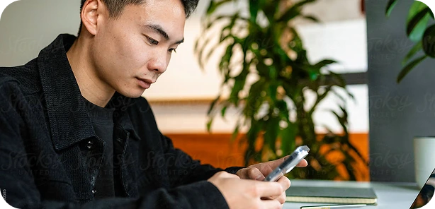 Young man in a black jacket sitting indoors, focused on his smartphone with a laptop and notebook on the table.