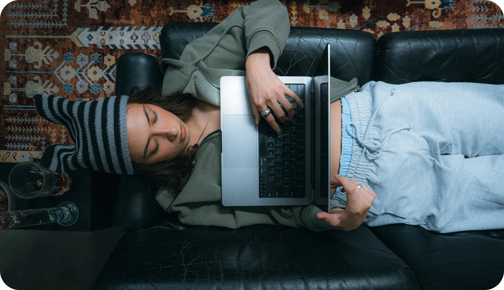 Person lying on a black leather couch with a laptop on their chest, wearing a striped beanie hat, green hoodie, and gray sweatpants.
