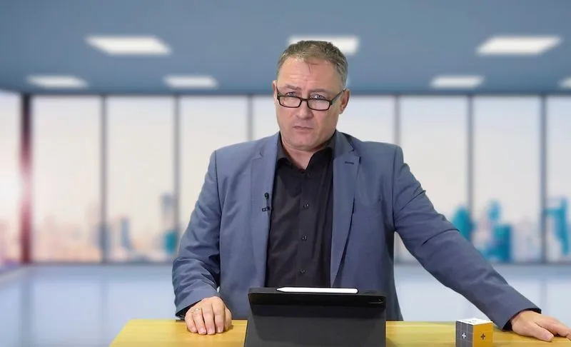 Ein Mann mit Brille trägt einen blauen Anzug und steht an einem Tisch in einem modernen Büro. Er sieht nachdenklich aus, während er auf ein Tablet schaut. Im Hintergrund sind hohe Fenster mit Blick auf eine Stadtsilhouette zu sehen.