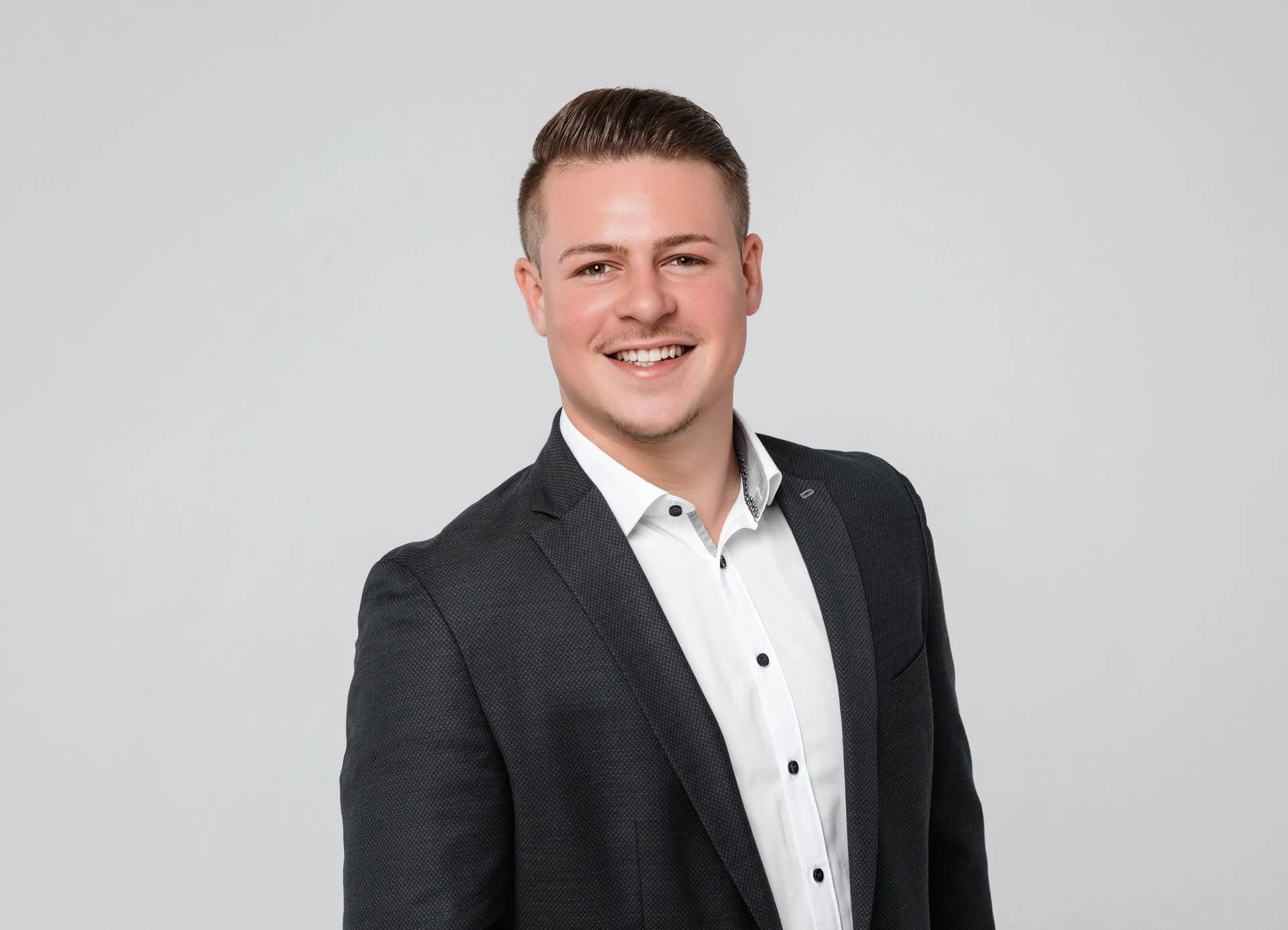 Ein lächelnder Mann in einem dunklen Anzug und einem weißen Hemd, der vor einem neutralen Hintergrund steht. Sein kurzes Haar ist ordentlich frisiert und er strahlt freundliche Zuversicht aus.