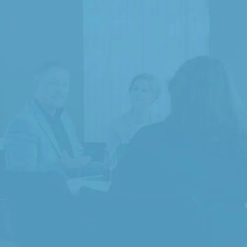 Drei Personen sitzen an einem Tisch in einem hellen Raum. Zwei Menschen unterhalten sich angeregt, während eine andere Person ihnen aufmerksam zuhört. Der Raum hat Lichtverhältnisse, die eine freundliche Atmosphäre schaffen.