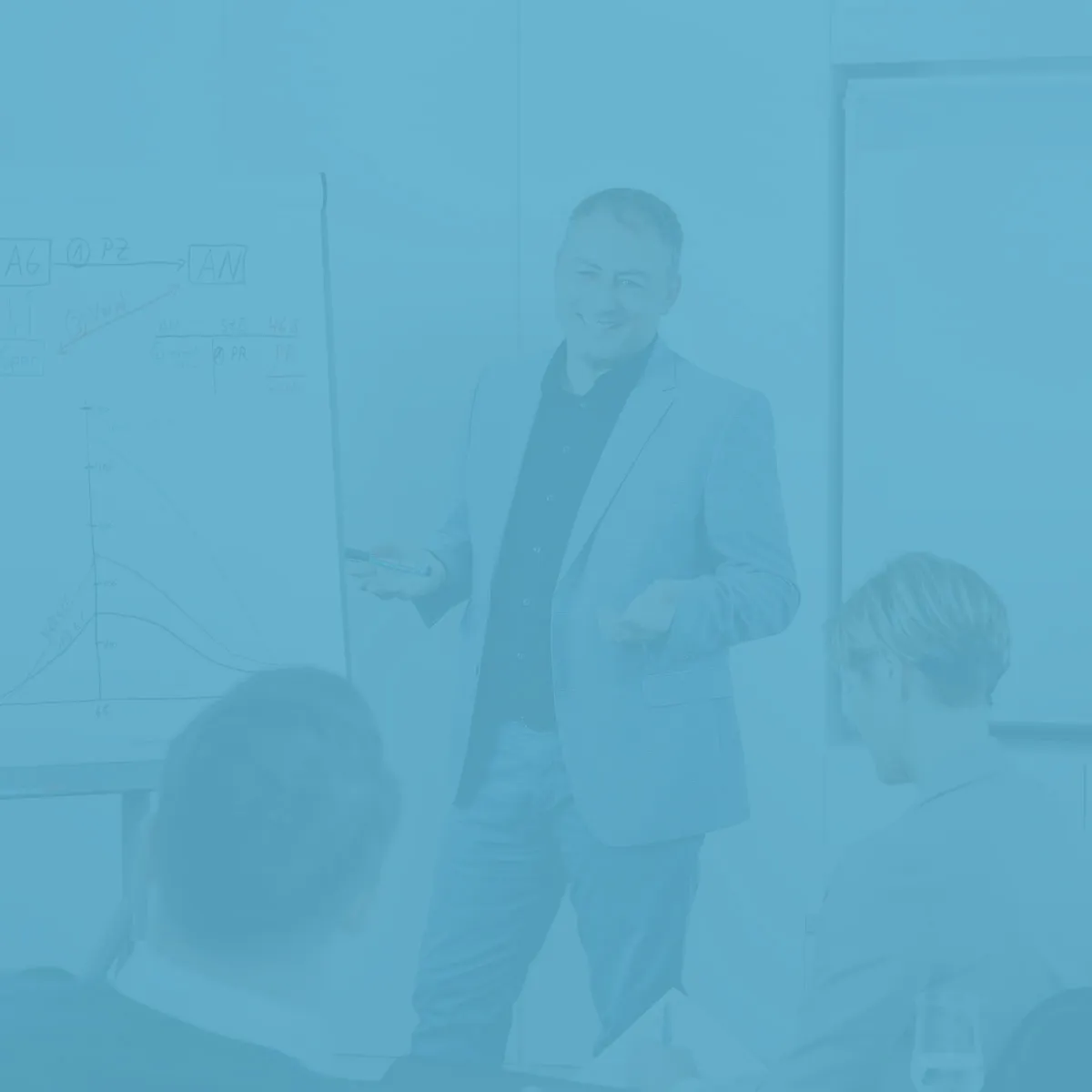 Ein Präsentator in einem grau-blauen Anzug spricht vor einem Flipchart während einer Besprechung, während das Publikum, bestehend aus mehreren Personen, ihm aufmerksam zuhört.