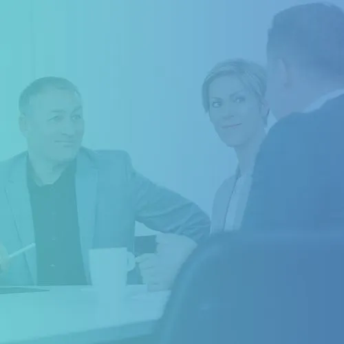 Drei Personen in einem geschäftlichen Meeting, die am Tisch sitzen. Ein Mann in einem grauen Anzug lächelt, während er auf einen Laptop zeigt. Eine Frau mit blonden Haaren schaut interessiert, während ein anderer Mann im Hintergrund spricht. Eine Tasse steht auf dem Tisch.