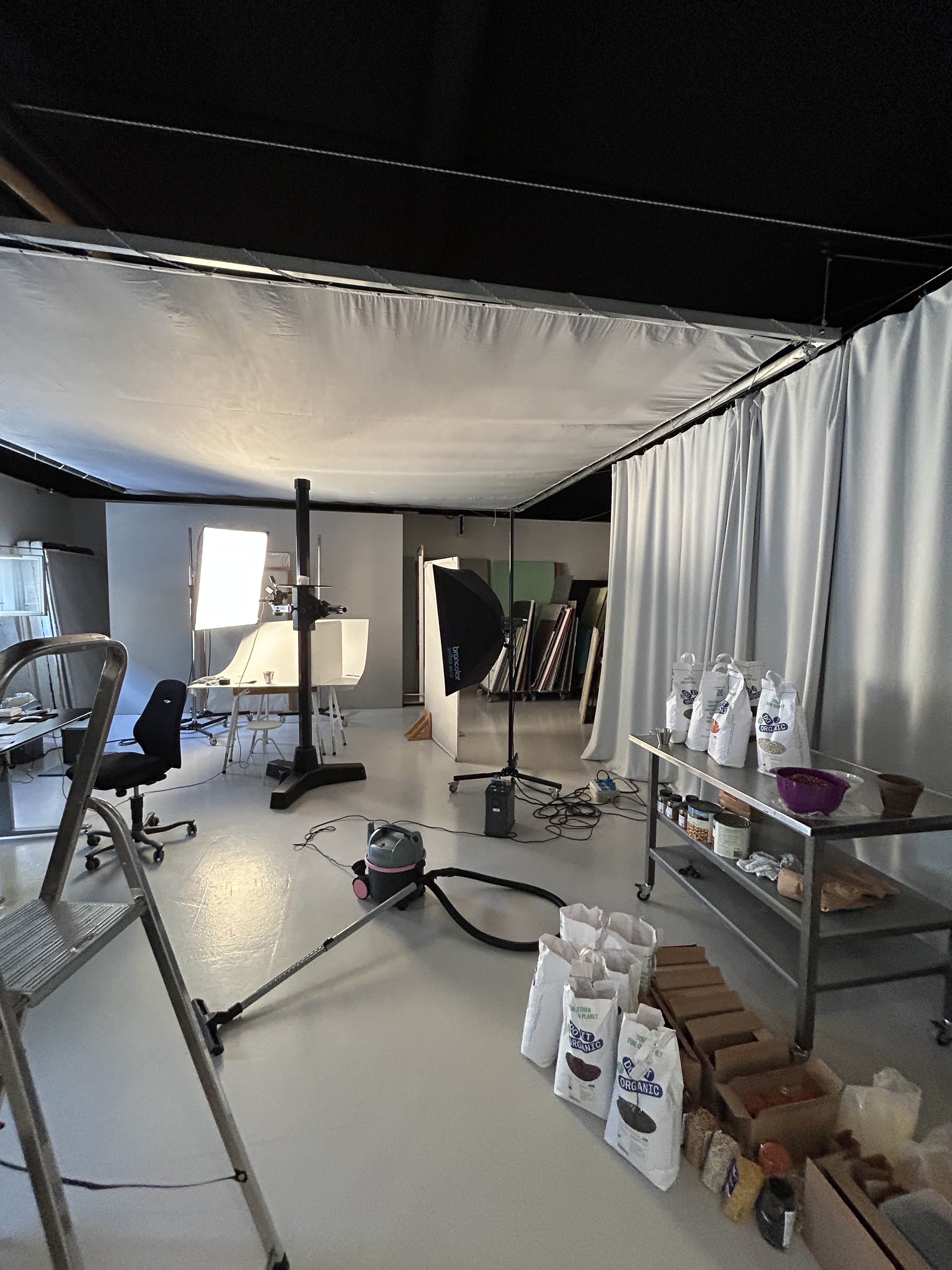 Fotostudie med lyspaneler, stole, en støvsuger, en trappestige og poser med økologiske varer på gulvet og en rullebord med beholdere.