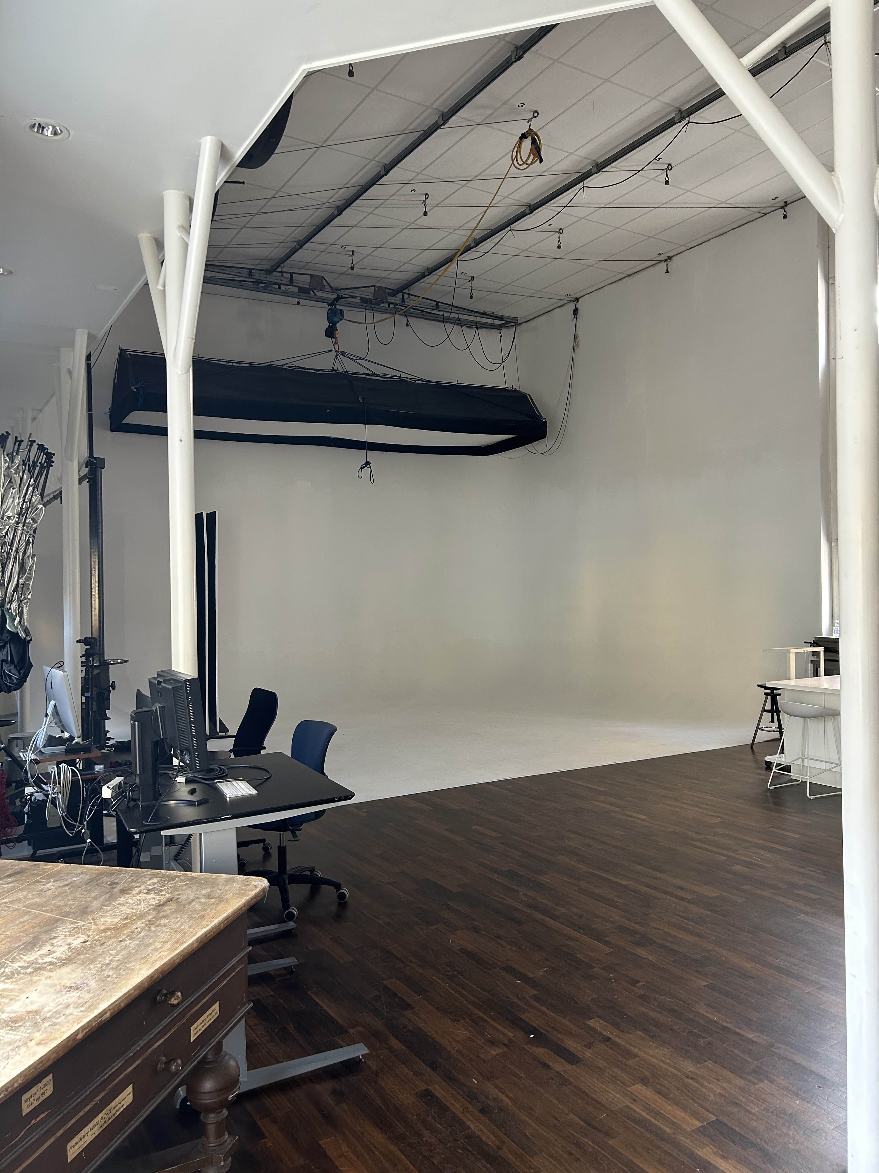 Et lyst fotosession-studie med mørkt trægulv, hvide vægge, kontorborde med computere og store belysningsudstyr ophængt i loftet.