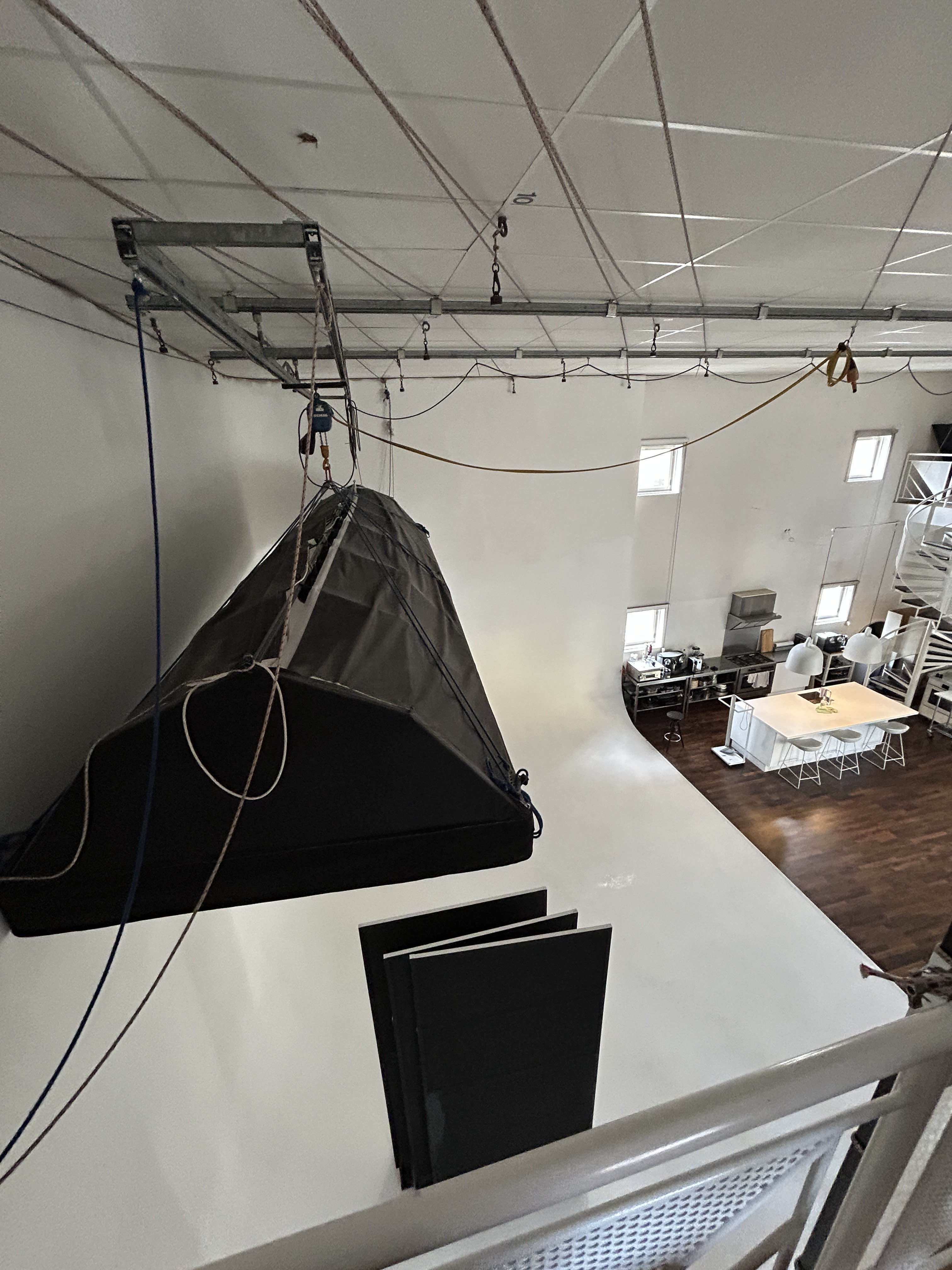 Indendørs fotostudie med hængende sort fotoudstyr og et lyst køkkenområde med bord og stole i baggrunden.