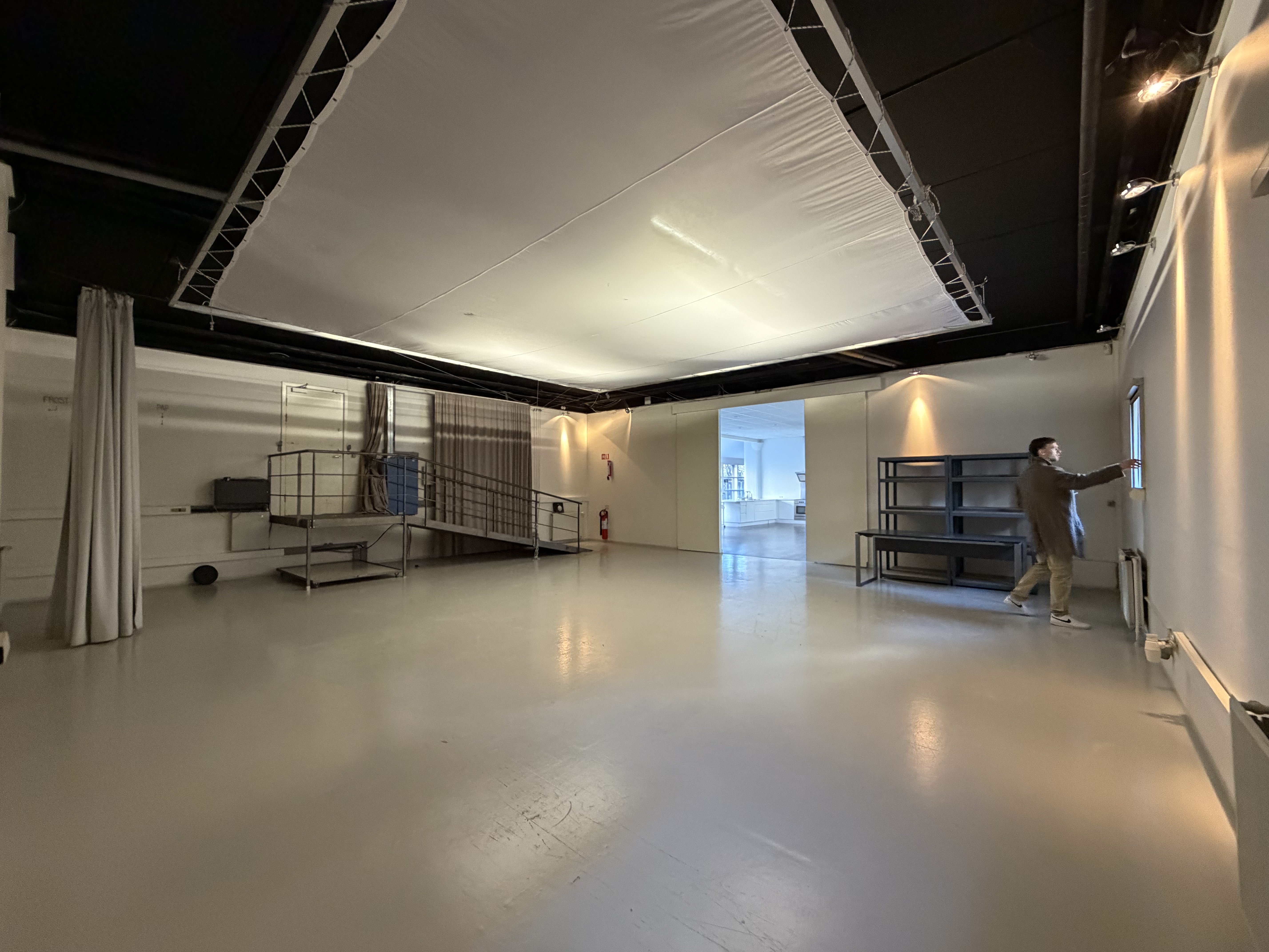 Stor tomt rum med lyst gulv og loftsophæng, en mand tjekker et vindue i højre side.