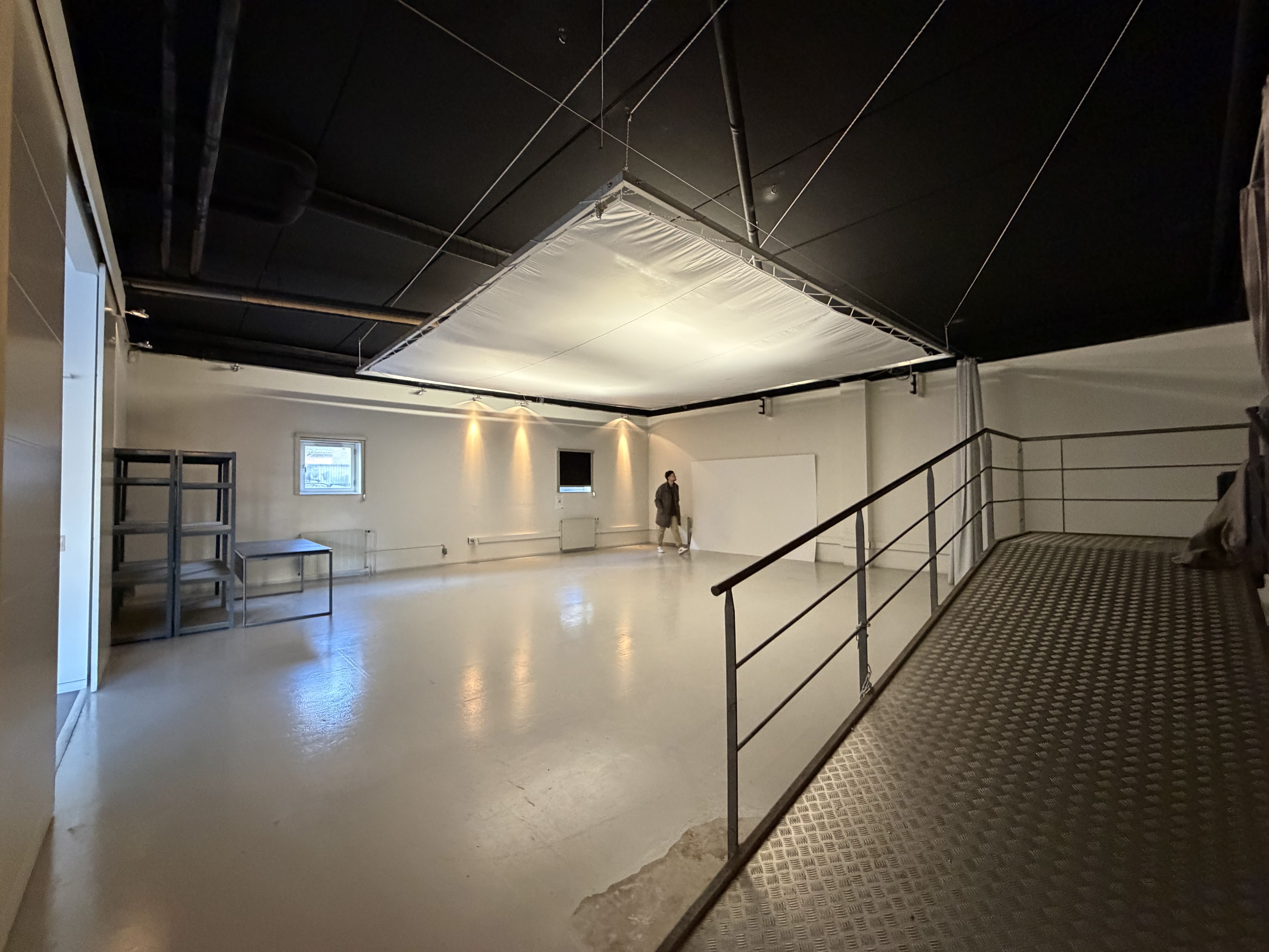 Stort, tomt rum med hvidt gulv, hvid væg og sort loft, en person står ved væggen, metalhylder og bord til venstre, rækværk og rampe til højre.