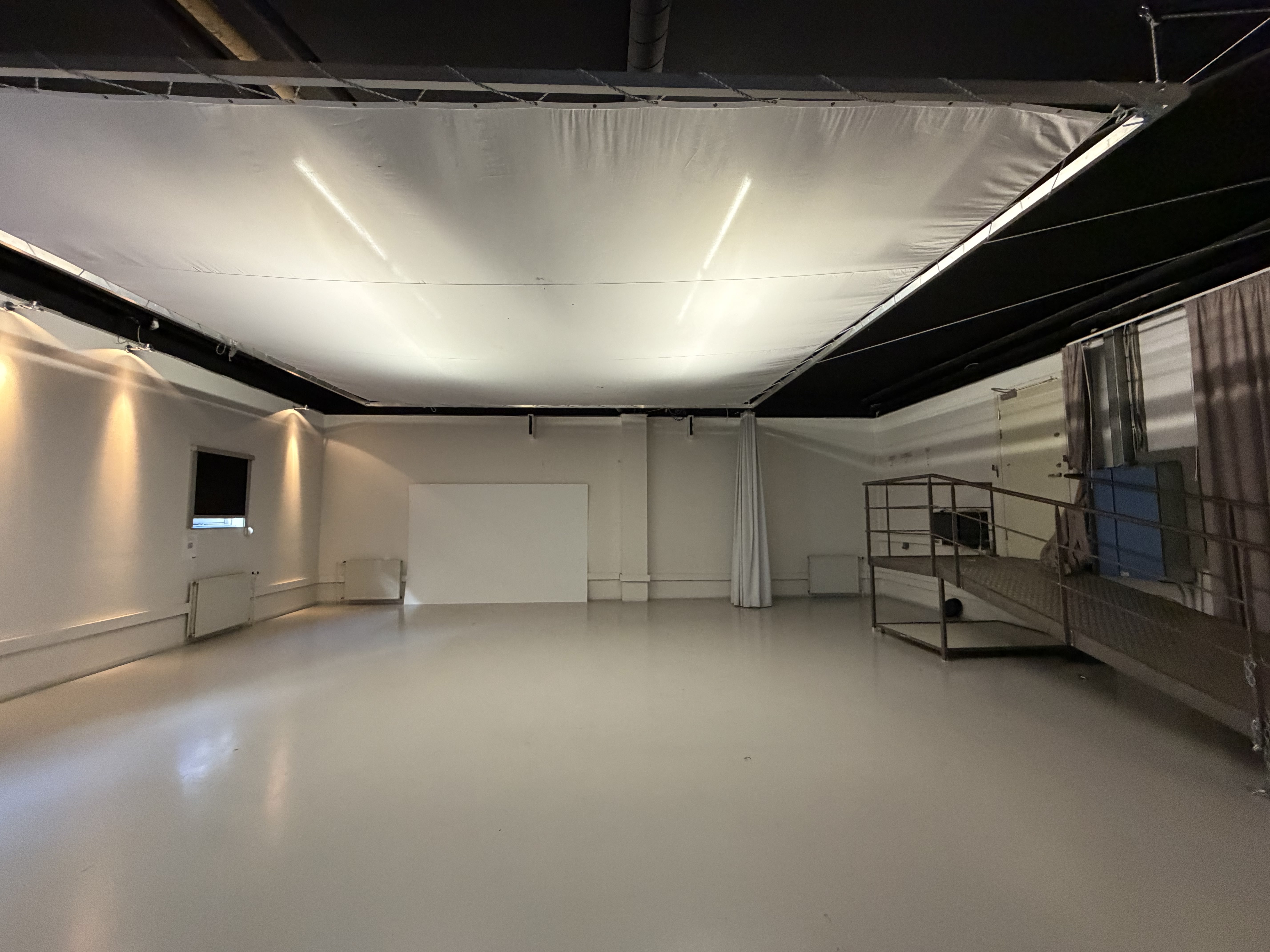 Stor tomt rum med hvidt gulv, hvide vægge, en hævet rampe til højre og en stor hvid skærm eller væg i baggrunden.