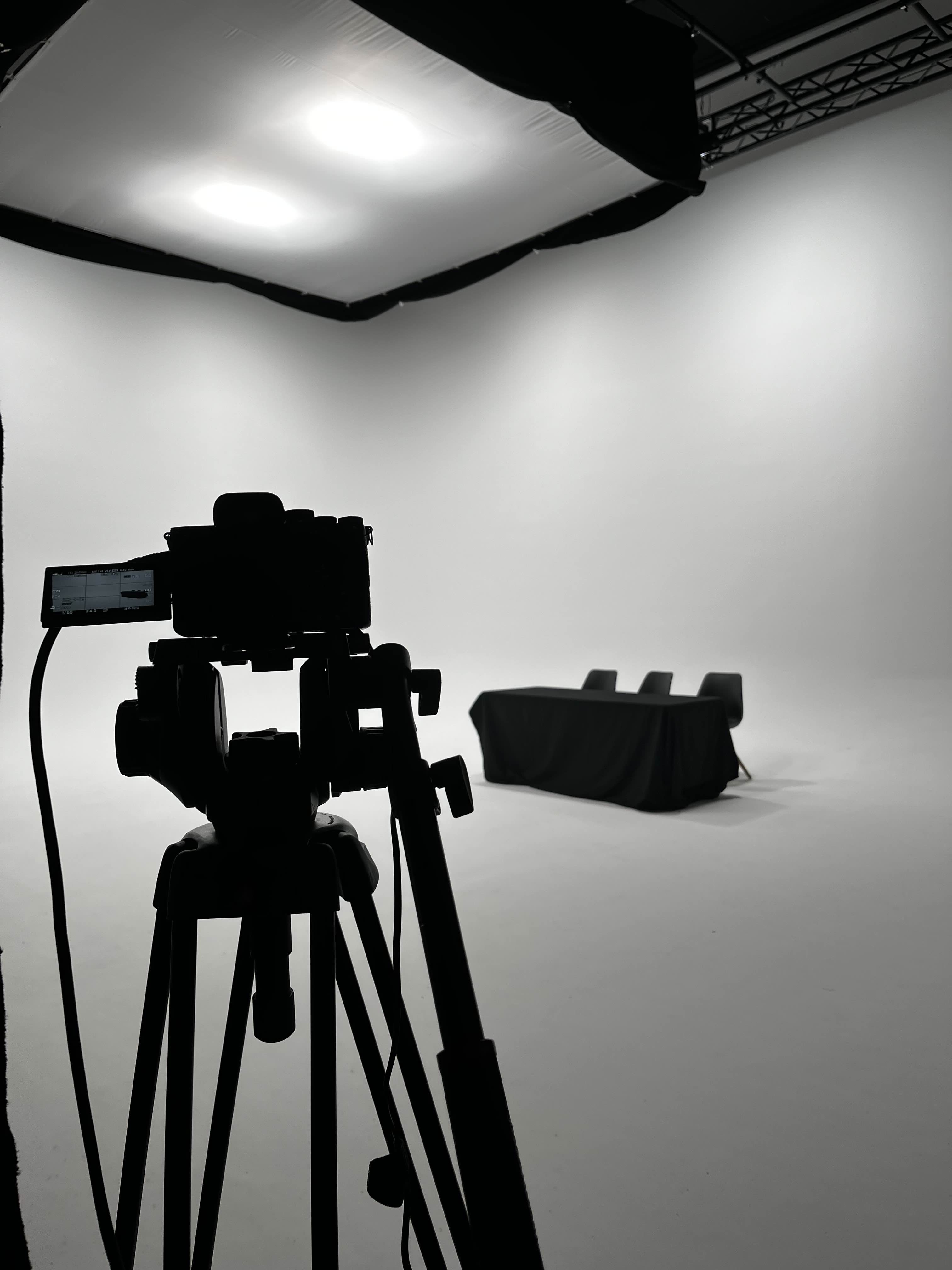 Silhuet af et kamera på stativ i et lyst fotostudie med et bord tildækket af sort stof og tre stole.
