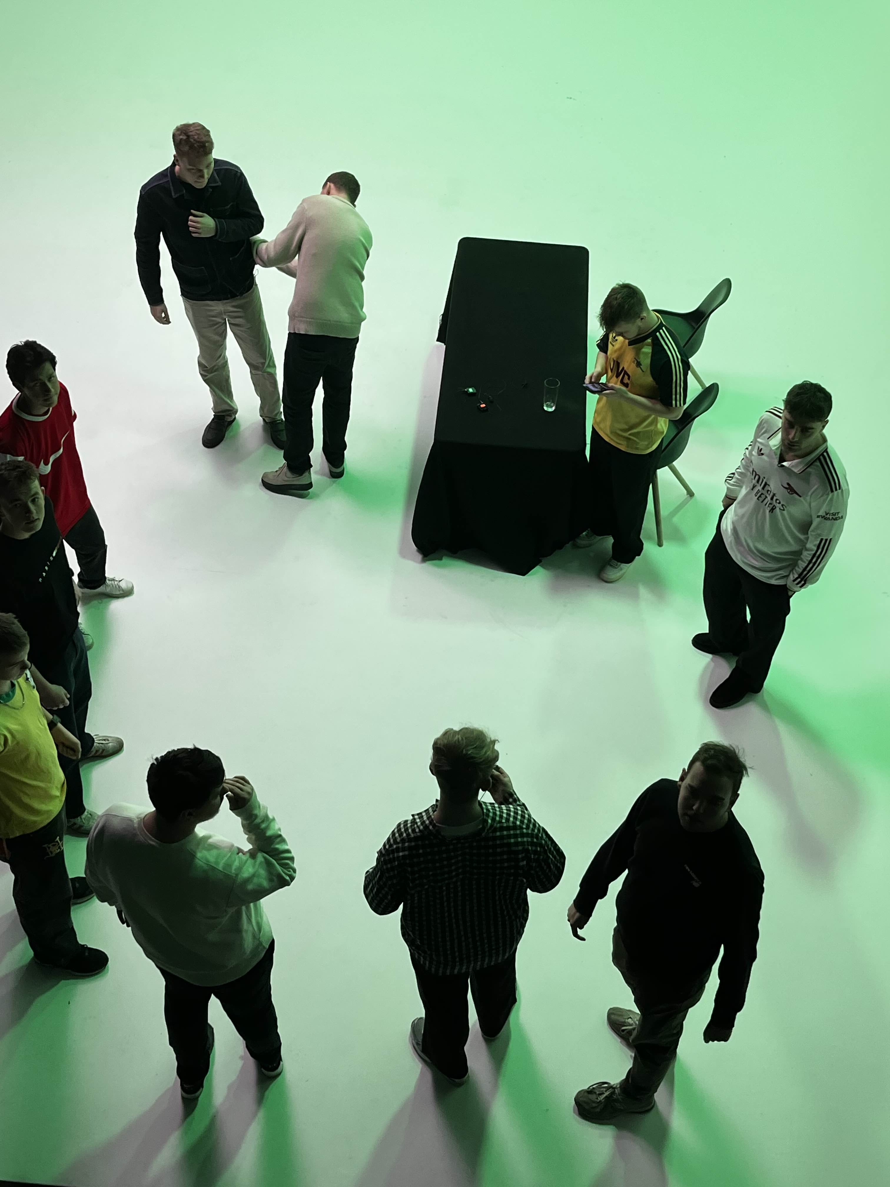 En gruppe unge mænd står spredt på et lyst gulv med grønt lys, omkring et bord med sort dug og to stole.