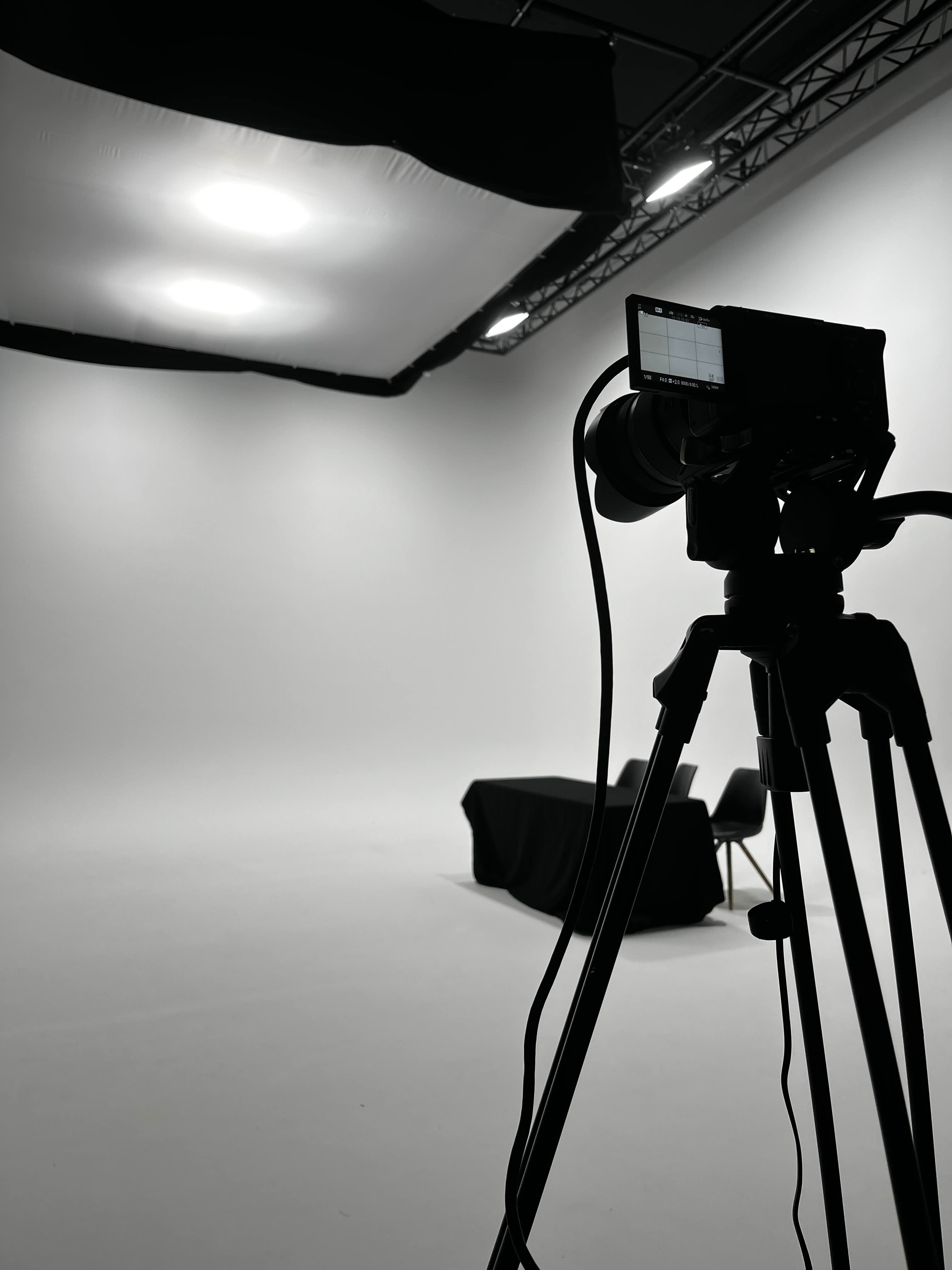 Silhuet af et professionelt videokamera på stativ i et lyst fotostudie med sortklædt bord og stol i baggrunden.