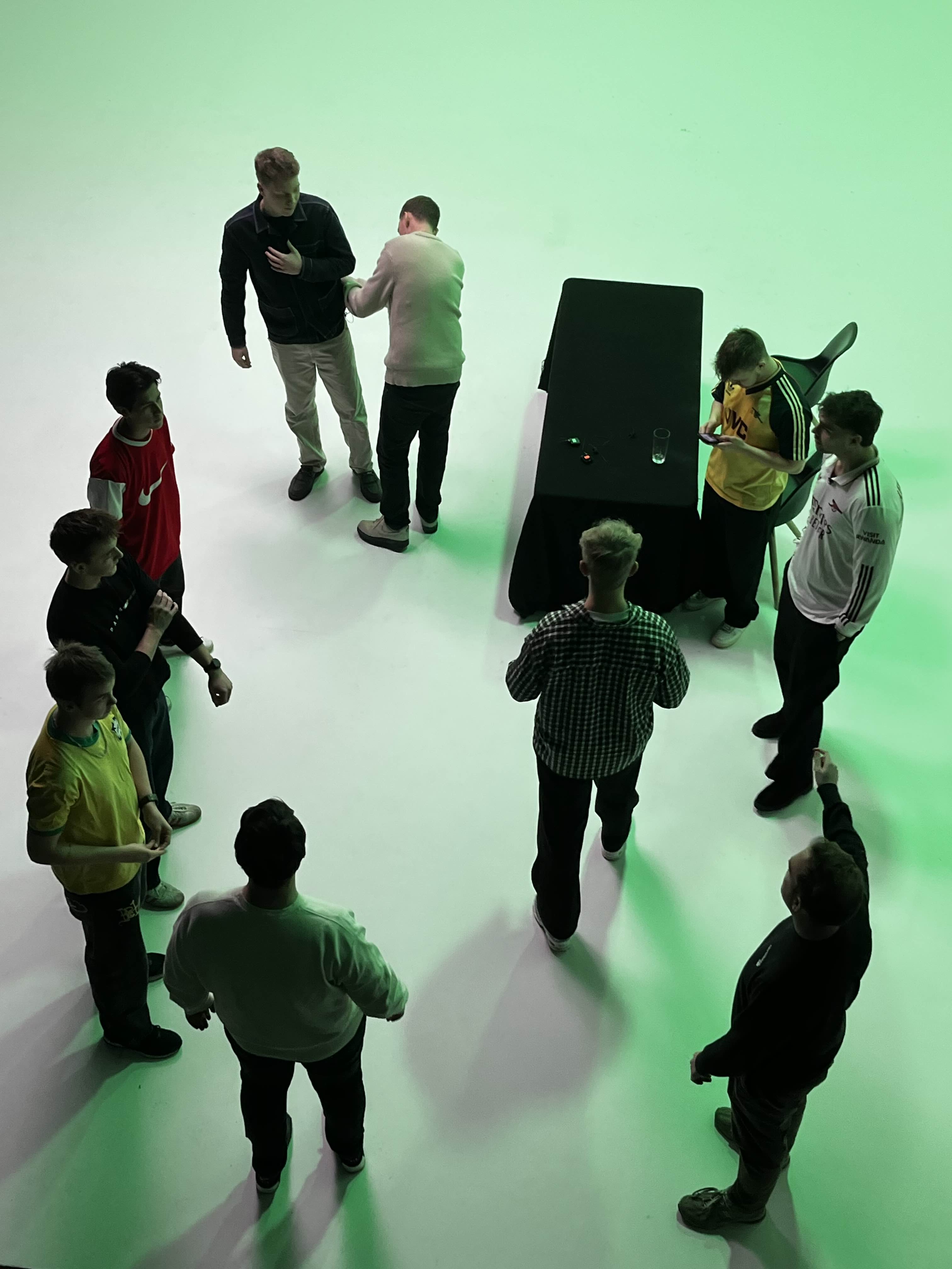 En gruppe unge mænd står i en cirkel på et lyst gulv med grøn belysning, nogle taler sammen, mens andre ser på deres telefon.