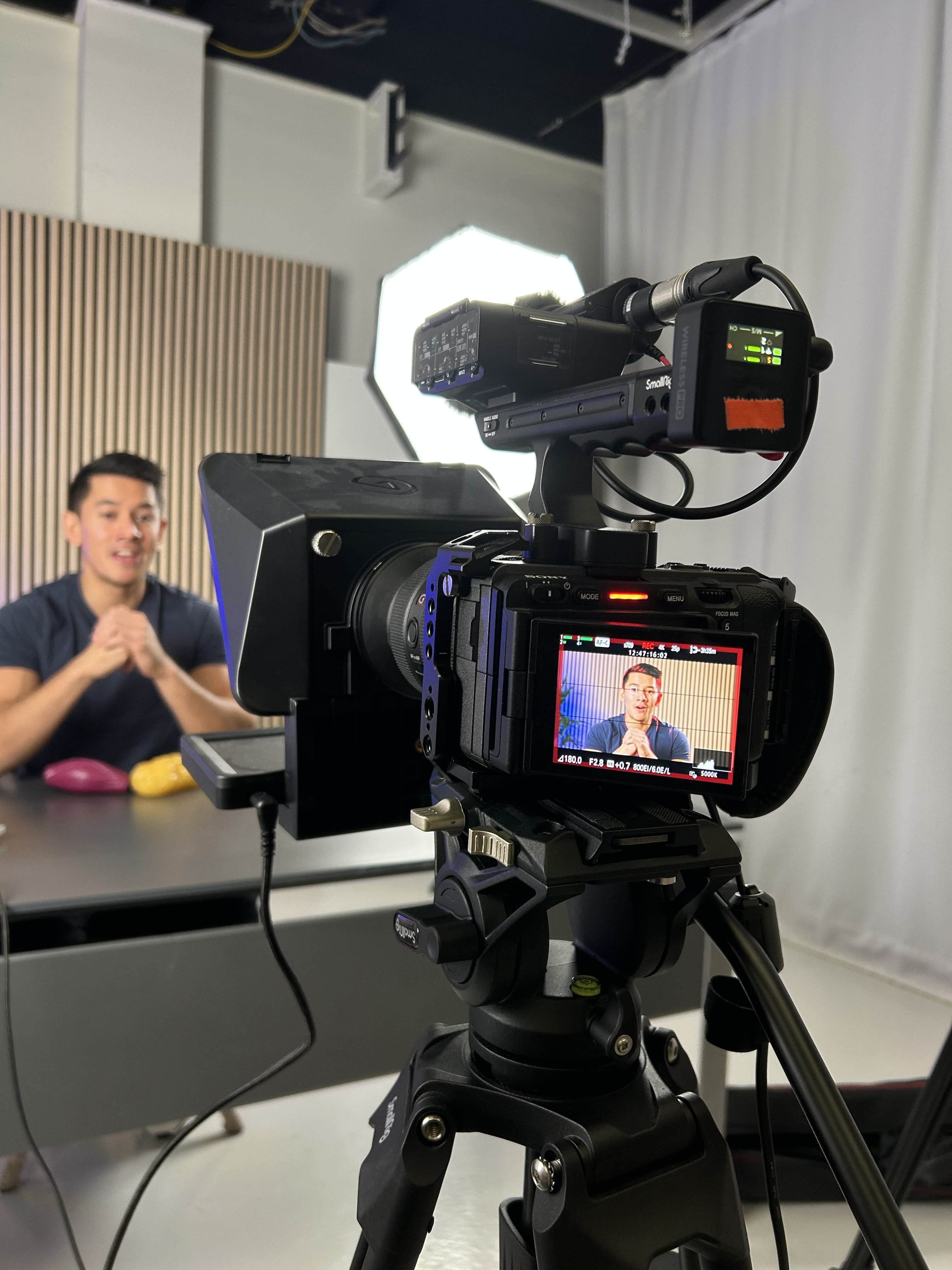 Professionelt videokamera på stativ optager en mand, der sidder ved et bord i et lyst studie.