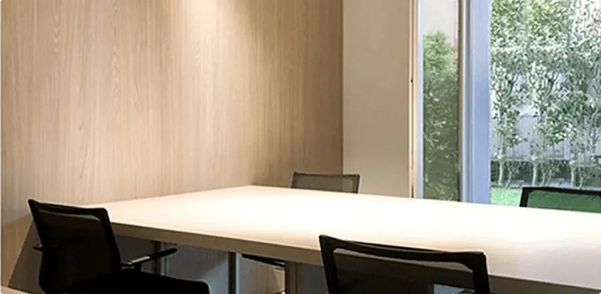 Sala riunioni minimalista con tavolo bianco rettangolare e quattro sedie nere, parete in legno chiaro e finestra con vista sul giardino.