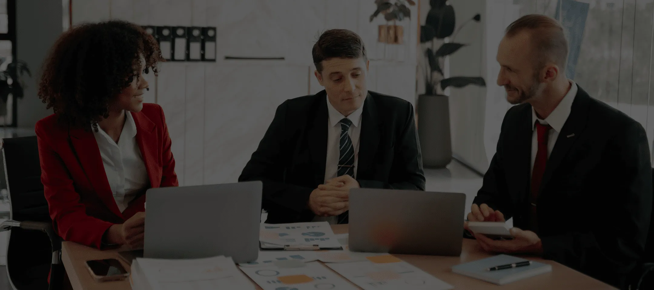 Tre professionisti in abito formale seduti a un tavolo con laptop e documenti, discutendo in un ufficio luminoso.