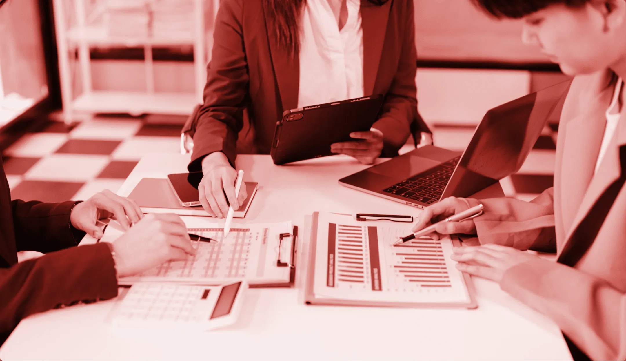 Tre persone in abiti formali che analizzano grafici e tabelle finanziarie su una scrivania con portatile e tablet.