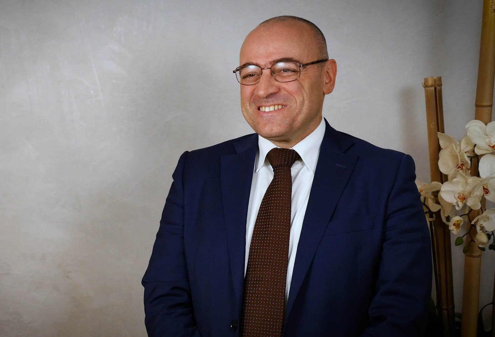 Uomo calvo con occhiali, vestito con giacca blu scuro, camicia bianca e cravatta marrone sorridente davanti a uno sfondo chiaro con fiori bianchi a destra.