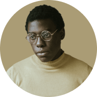 Portrait of a young man with short curly hair wearing round glasses and a beige turtleneck sweater against a muted beige background.