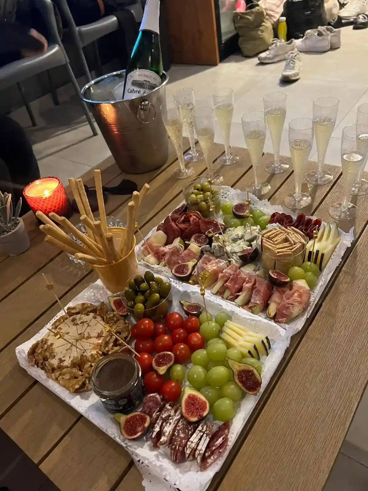 Wooden table set with two trays of assorted appetizers including grapes, figs, cherry tomatoes, crackers, cheeses, cured meats, olives, and breadsticks, alongside champagne glasses and a bottle of sparkling wine in an ice bucket.