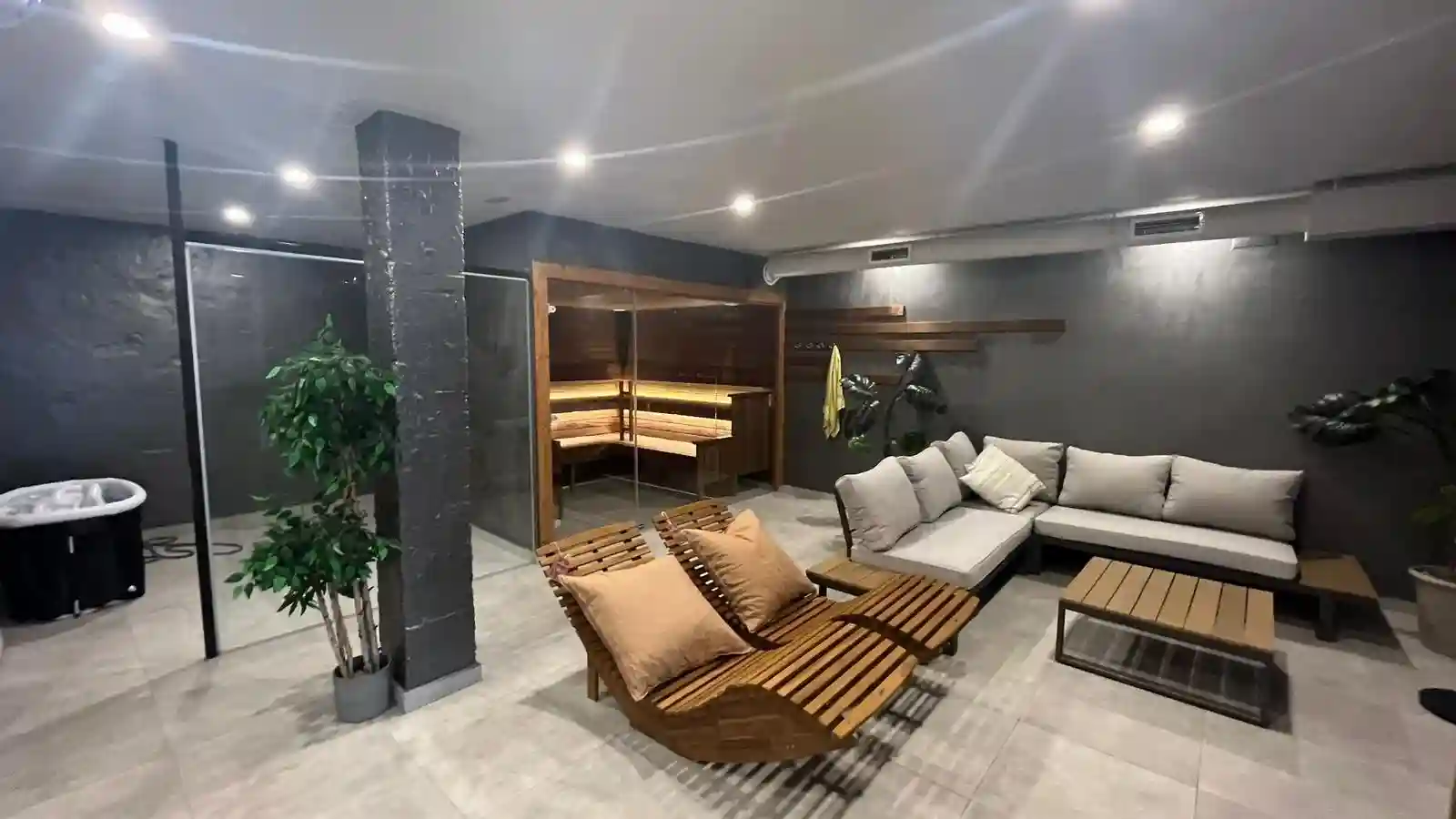 Modern relaxation room with a wooden sauna, gray sectional sofa, wooden lounge chairs with cushions, and potted plants.