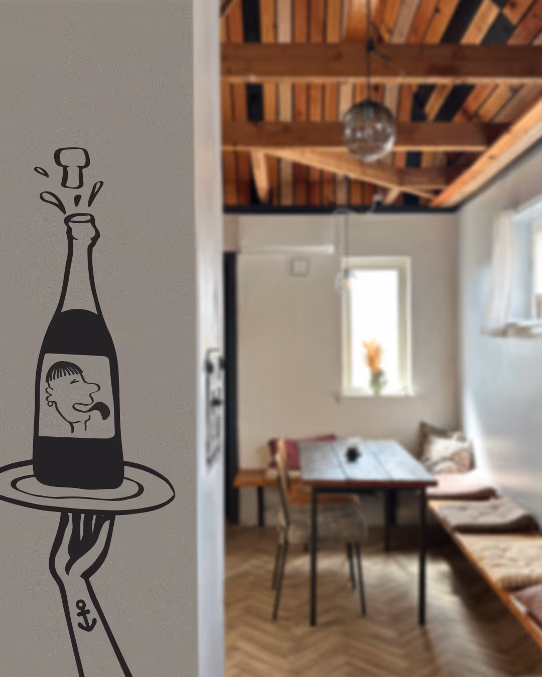 Vægmaling af en arm med anker-tatovering, der holder et fad med en boblende champagneflaske med en tegnet etiket, i et hyggeligt rum med træloft og spisebord.