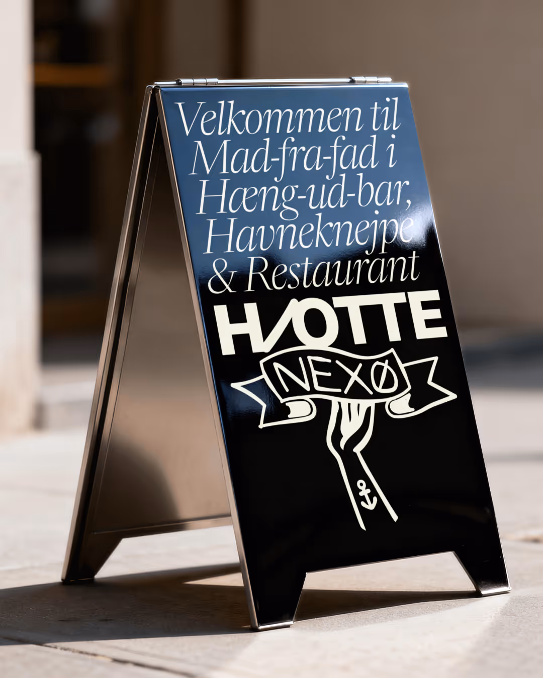 Sort gadestander med hvid tekst, der byder velkommen til mad-fra-fad i Hæng-ud-bar, Havneknejpe & Restaurant HJØTTE NEXØ.