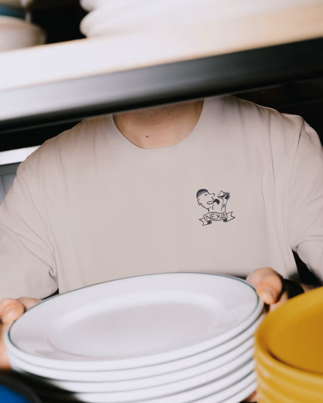 Person der bærer en stak hvide tallerkener med en t-shirt, der har en lille tegning af en person, der drikker fra en flaske med teksten 'NEXØ'.