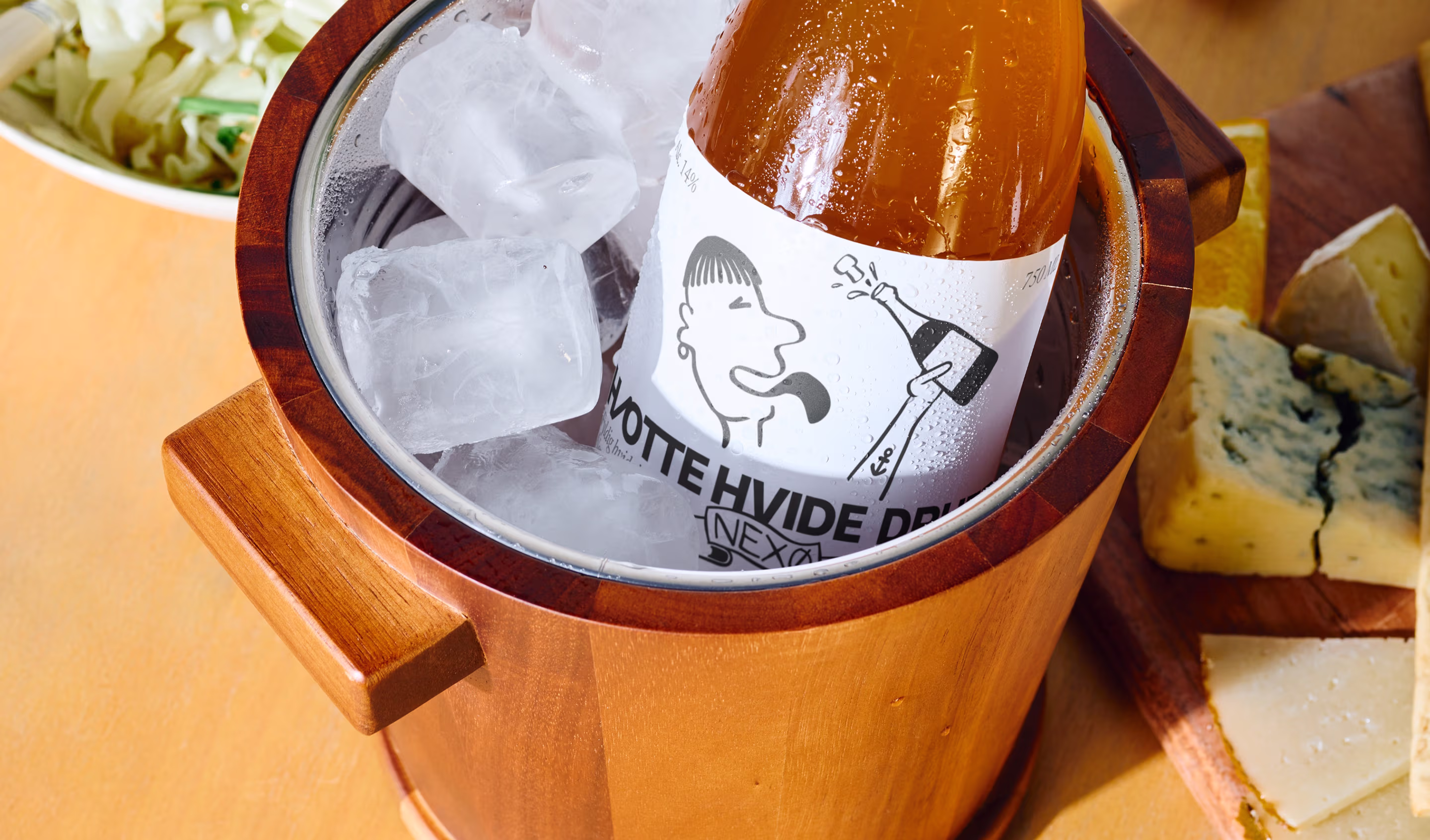 Flaske med drik i en trækøler med is, ved siden af forskellige oste på et skærebræt.