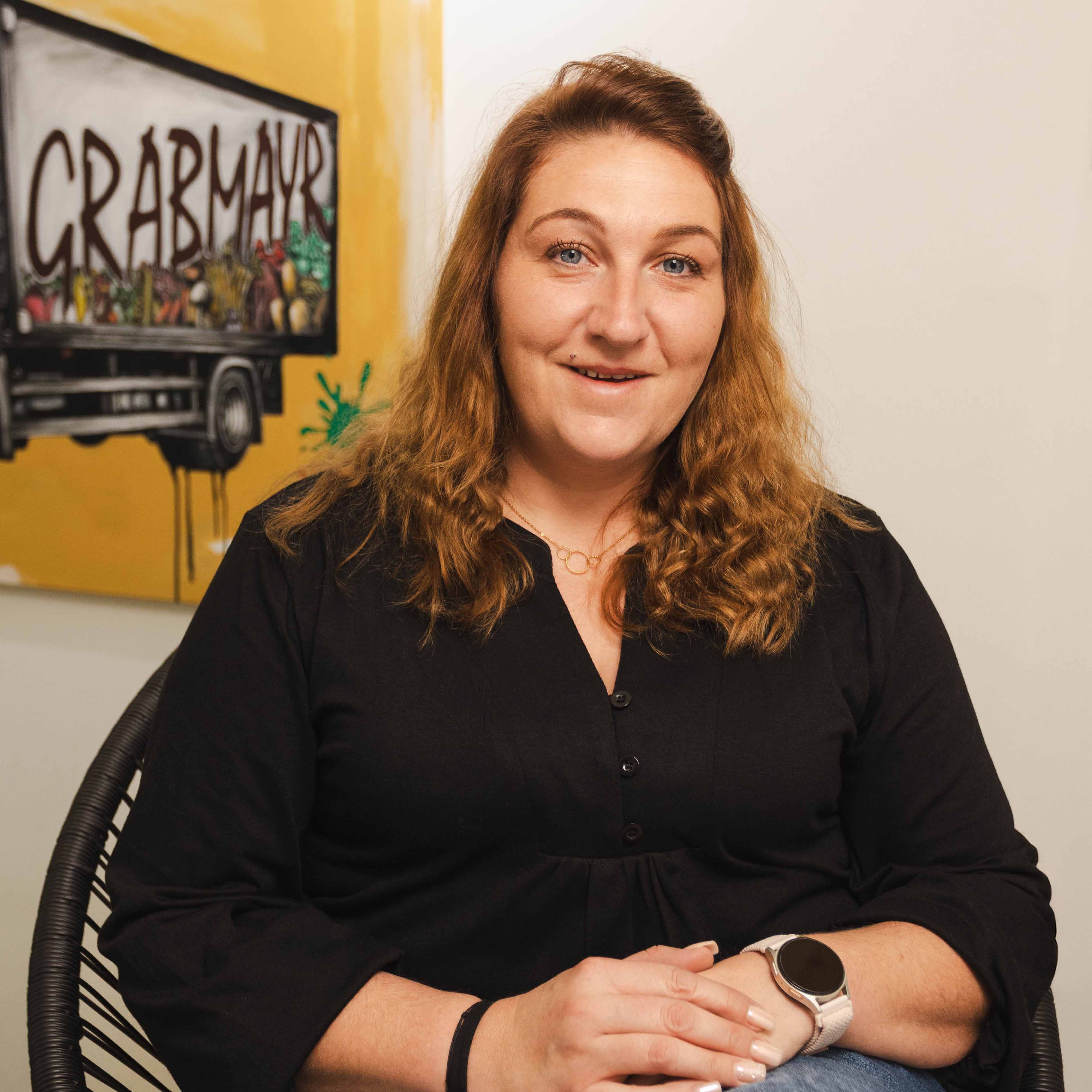 Frau mit lockigem braunem Haar trägt schwarzen Pullover und Armbanduhr, sitzt lächelnd vor gelbem Wandbild mit Schriftzug ‚GRABMAYR‘.