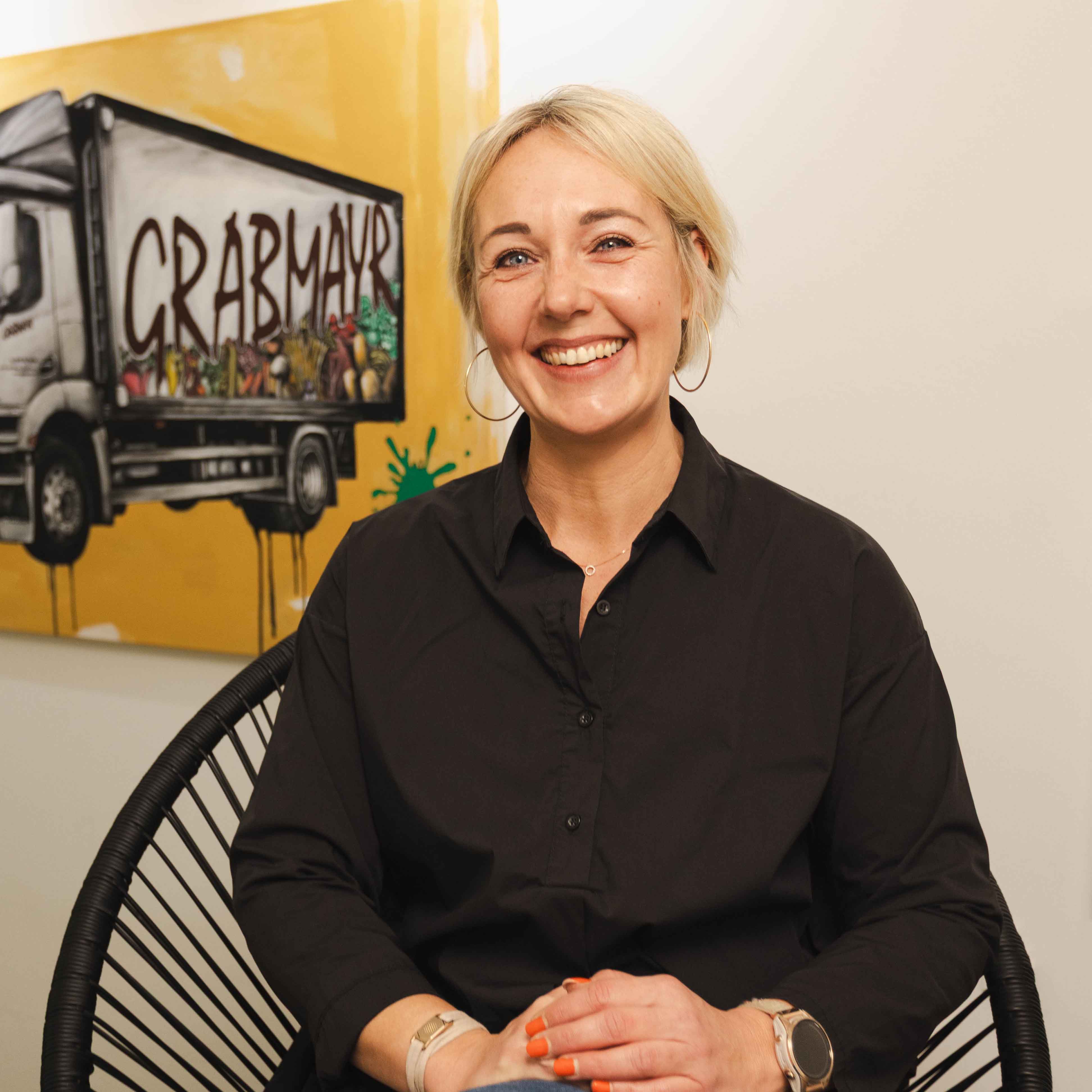 Lächelnde blonde Frau mit kurzem Haar und orange lackierten Fingernägeln sitzt auf einem schwarzen Stuhl vor einer Wand mit einem gelben Gemälde eines Lkw.