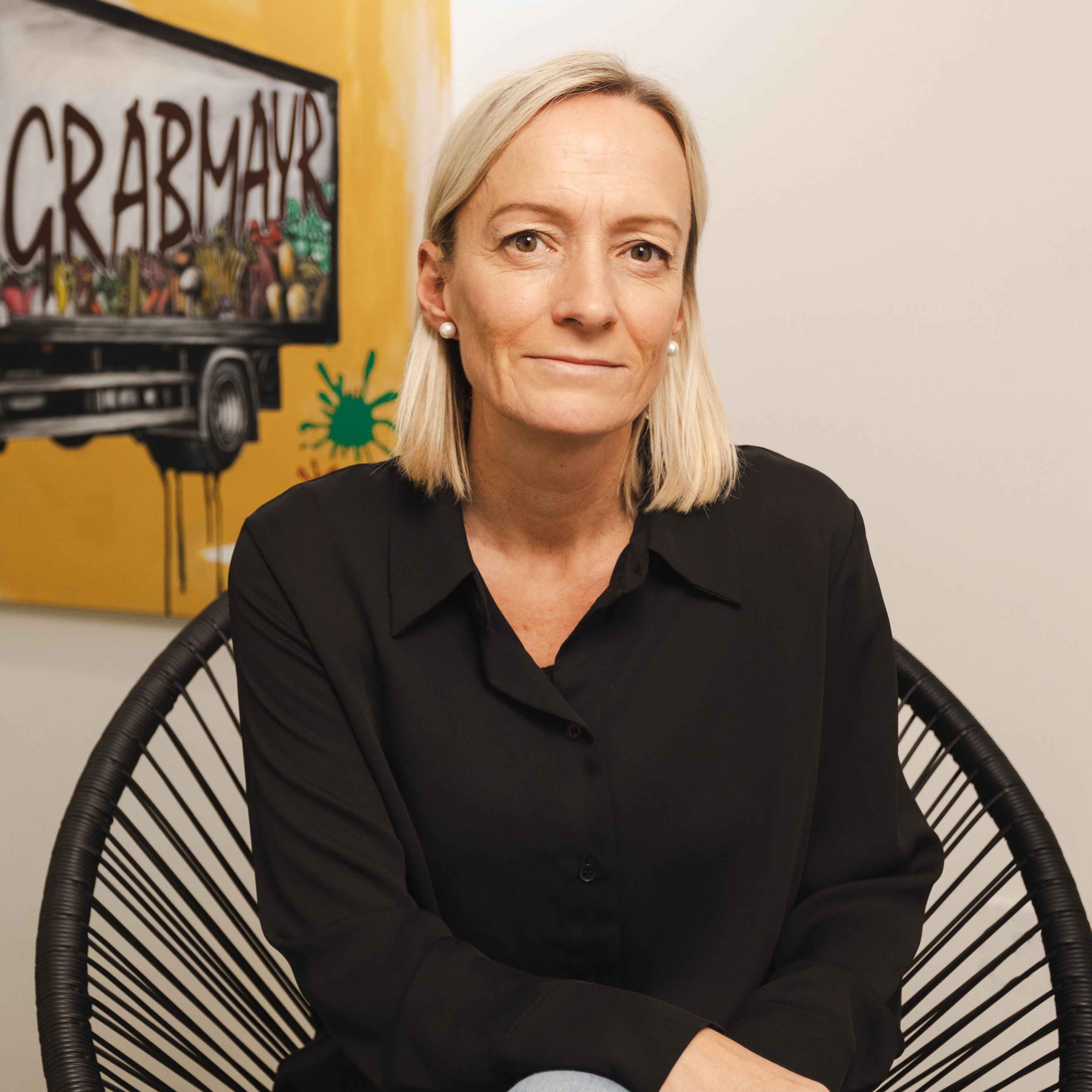 Frau mit kurzen blonden Haaren und schwarzer Bluse sitzt vor einer Wand mit einem Bild eines Lastwagens und dem Schriftzug Grabmayr.