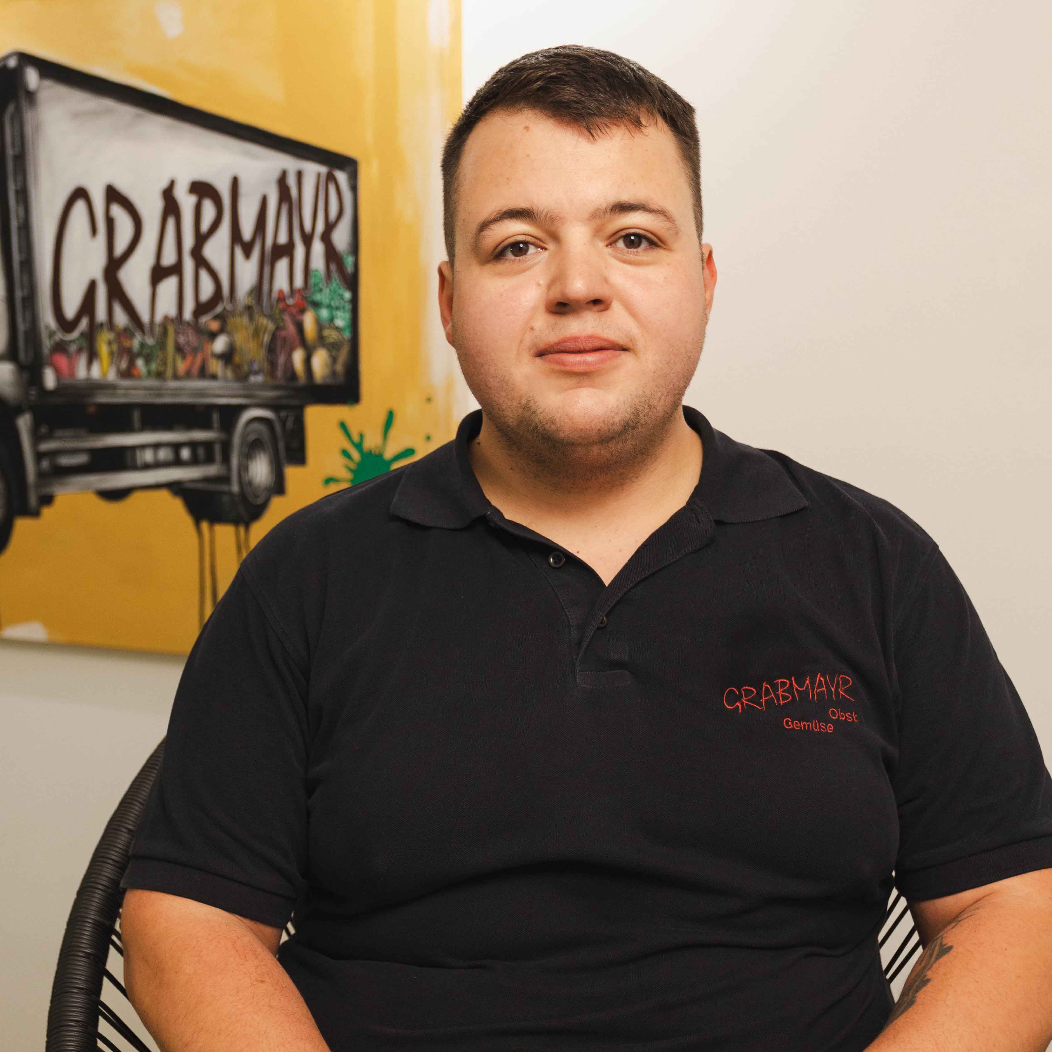 Porträt eines Mannes mit schwarzem Poloshirt mit der Aufschrift ‚Grabmayr Obst Gemüse‘ vor einer Wand mit einem Gemälde eines Lastwagens mit ‚Grabmayr‘-Schriftzug.