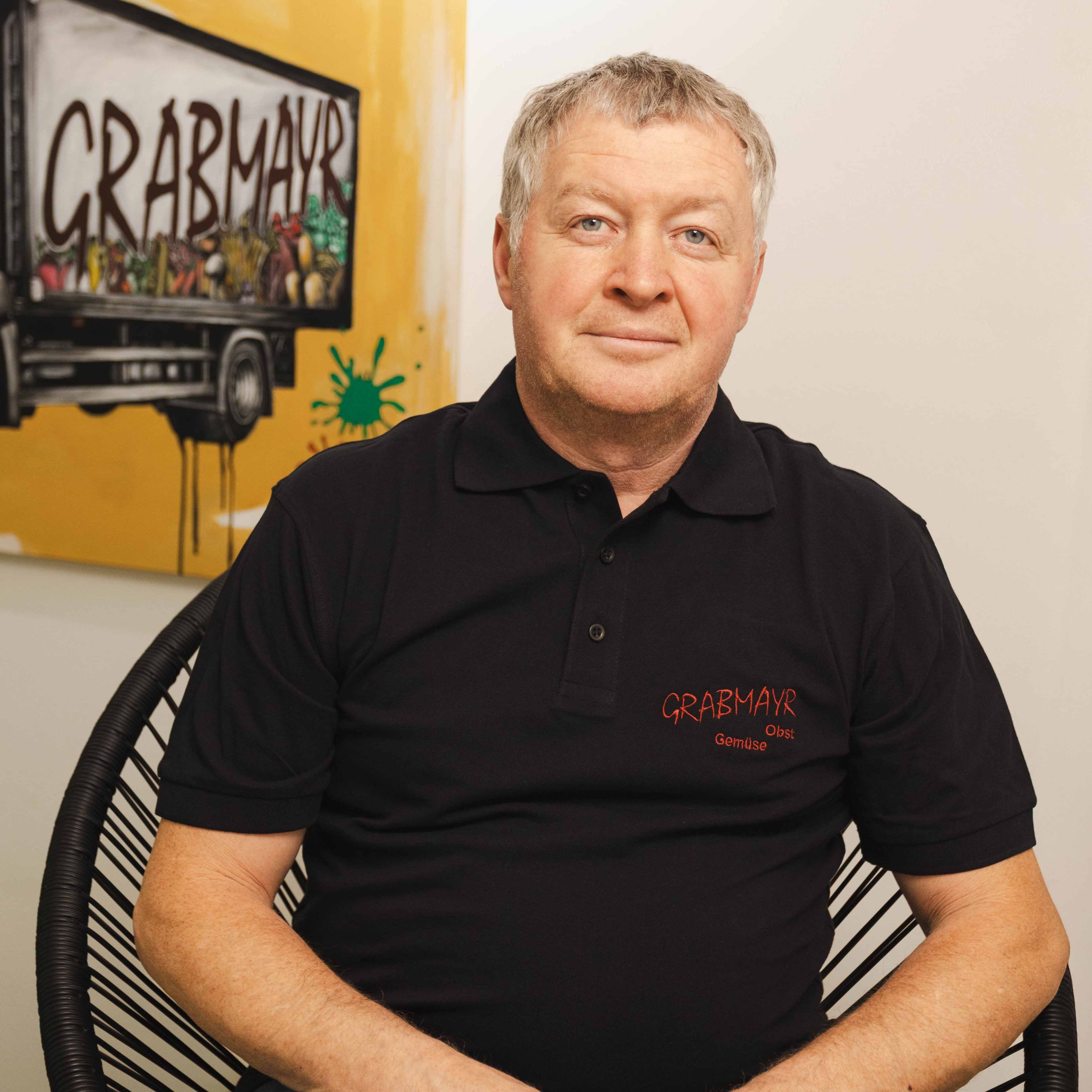 Mittelalter Mann mit blondem Haar in einem schwarzen Polohemd mit der Aufschrift ‚GRABMAYR Obst Gemüse‘ sitzt vor einer gelben Wand mit einem Wandbild eines Lastwagens.