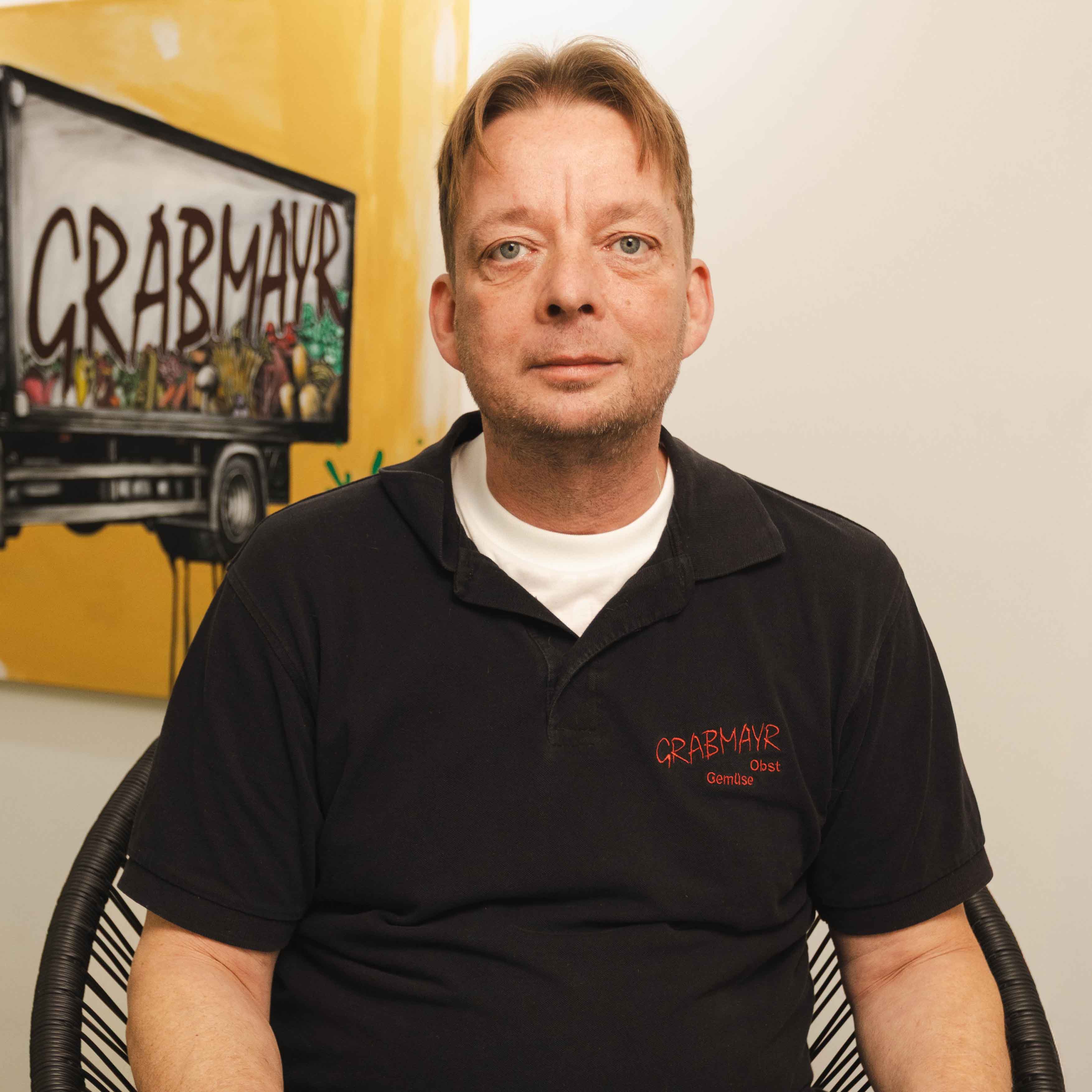 Mann mit schwarzem Polohemd mit der Aufschrift „GRABMAYR Obst Gemüse“ sitzt vor einer Wand mit einem Bild eines Lastwagens mit dem Schriftzug GRABMAYR.
