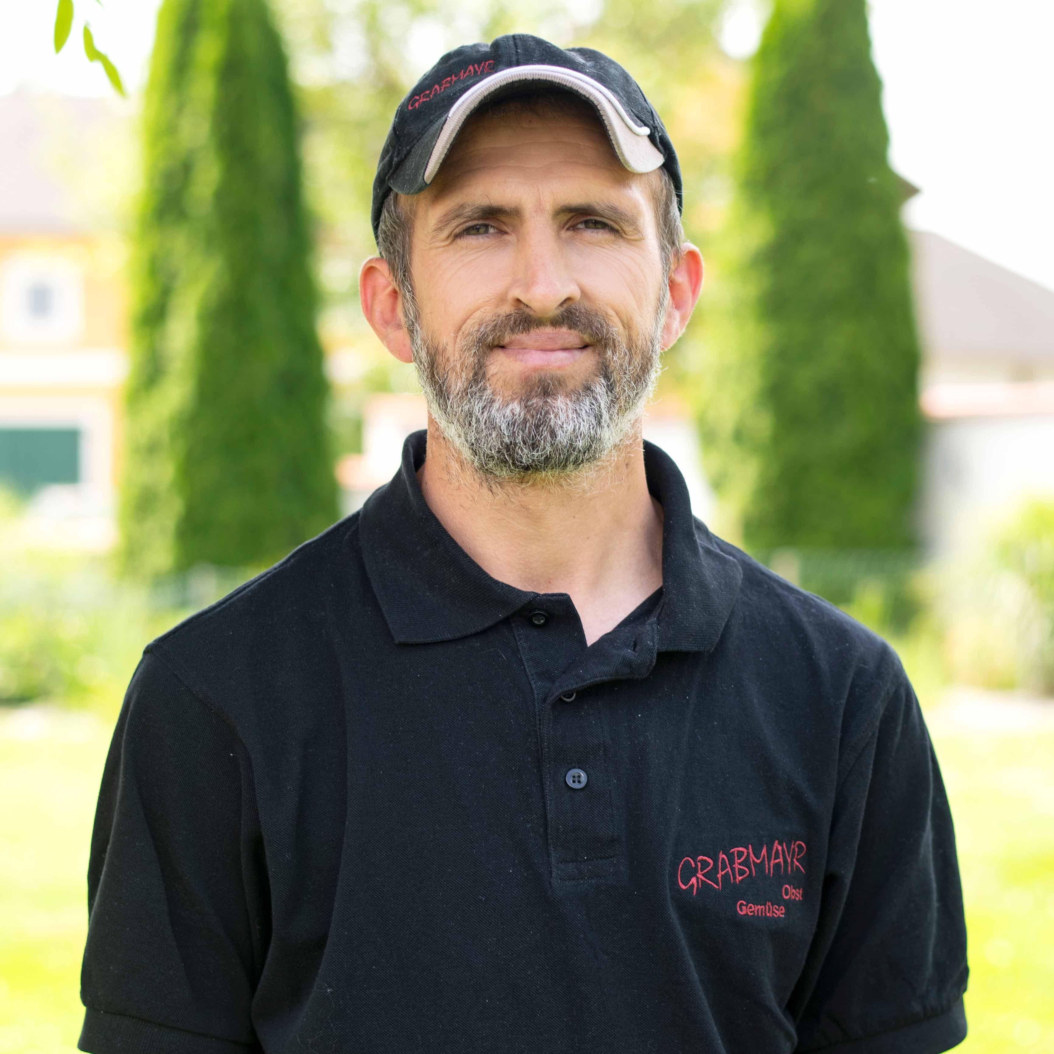 Mann mit grauem Bart und schwarzer Kappe trägt schwarzes Polohemd mit rotem Schriftzug Grabmayr Obst Gemüse, steht im Freien.