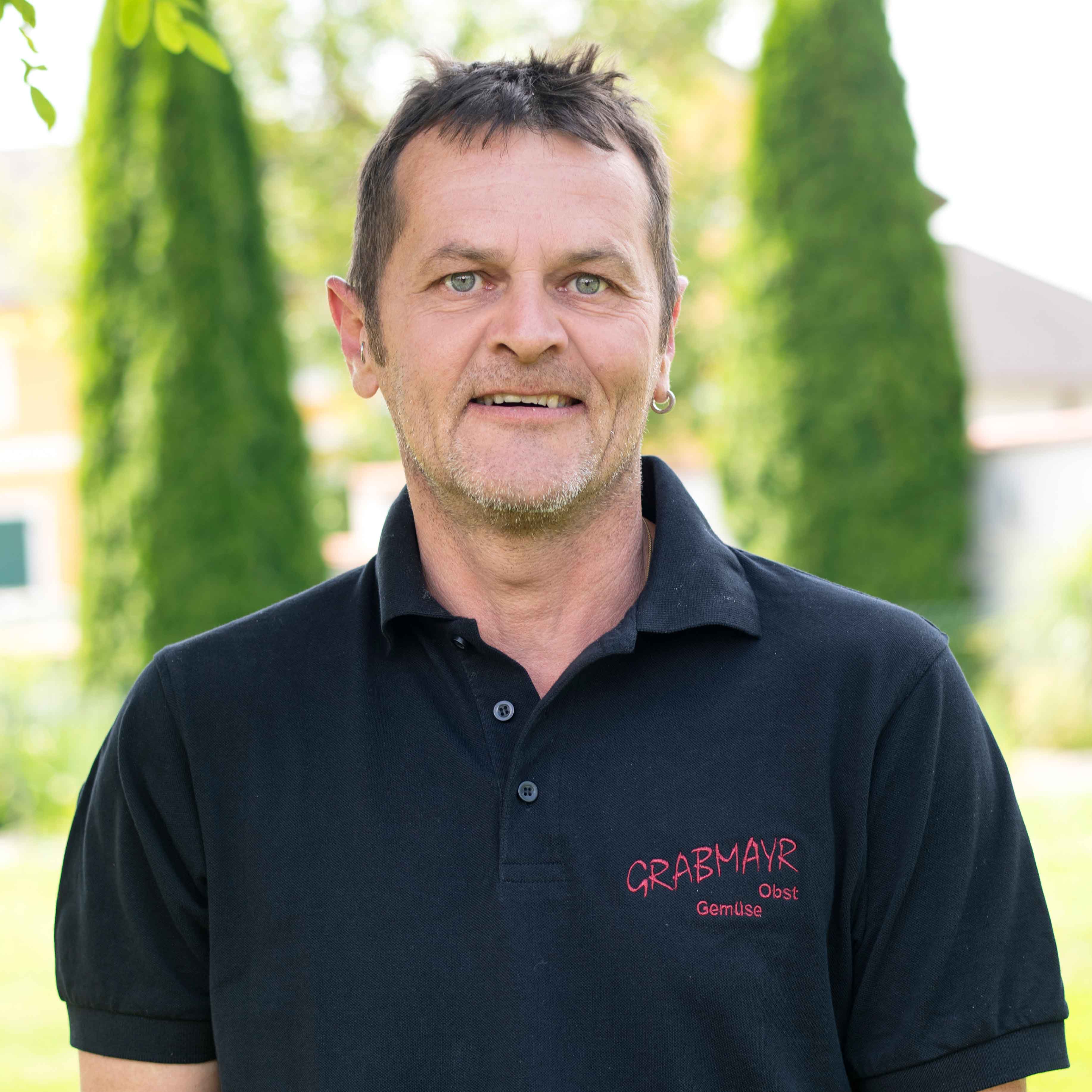 Mann mit kurzen dunklen Haaren und Bart trägt schwarzes Polohemd mit Aufschrift 'GRABMAYR Obst Gemüse' vor grünem Hintergrund.