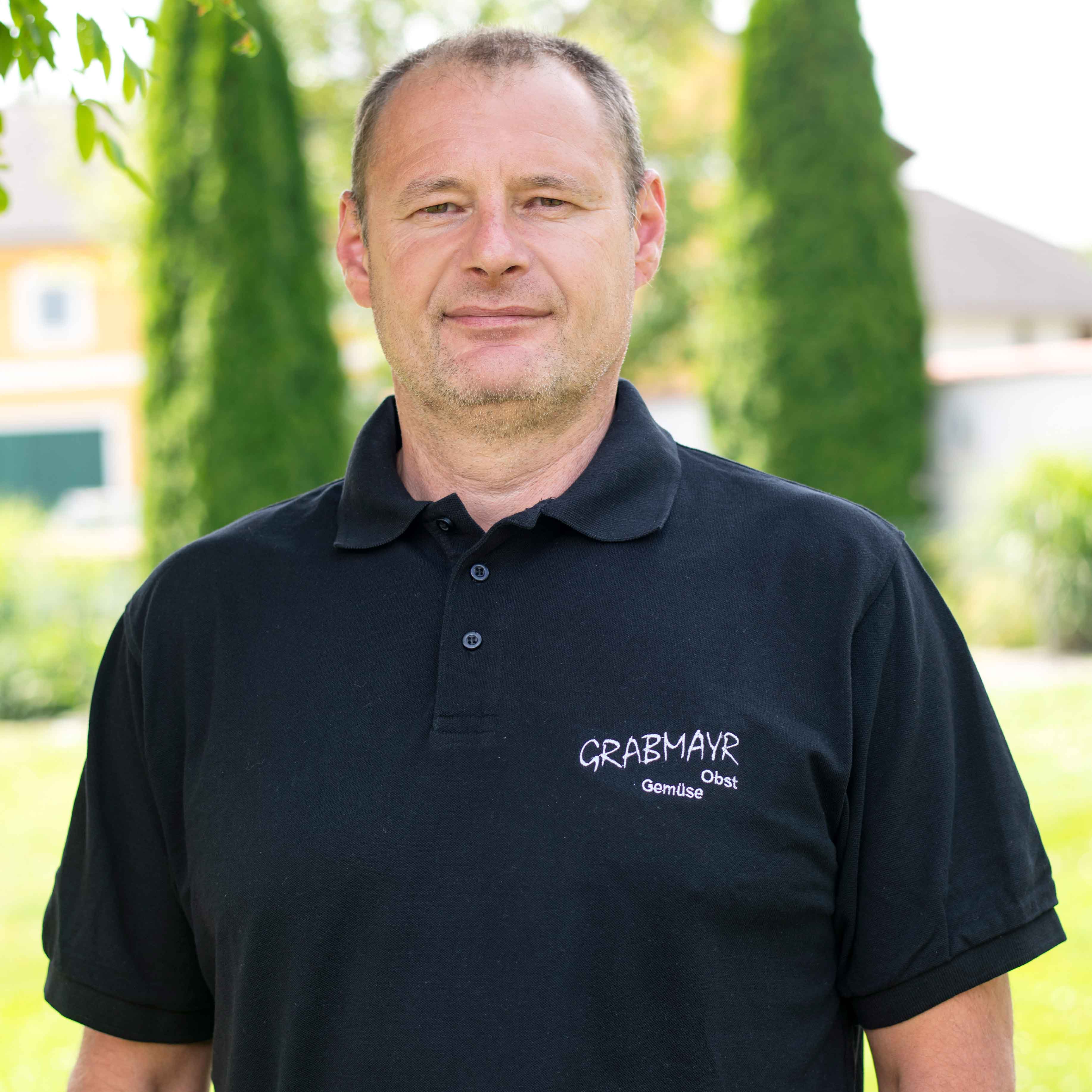 Mann trägt schwarzes Polohemd mit Aufschrift ‚GRABMAYR Obst Gemüse‘ vor grüner Gartenlandschaft.