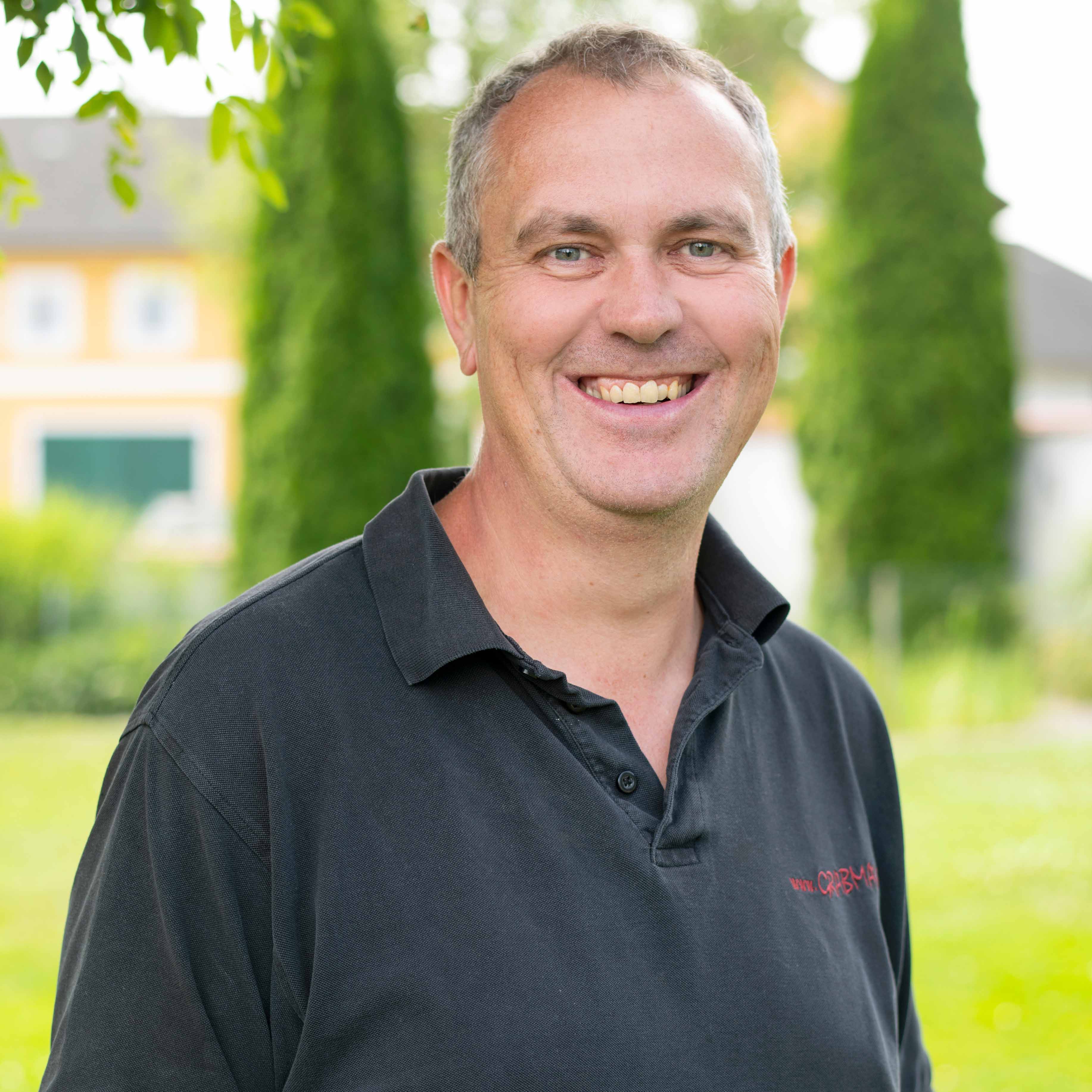Lächelnder Mann in schwarzem Poloshirt vor einem Garten mit Bäumen und Häusern im Hintergrund.