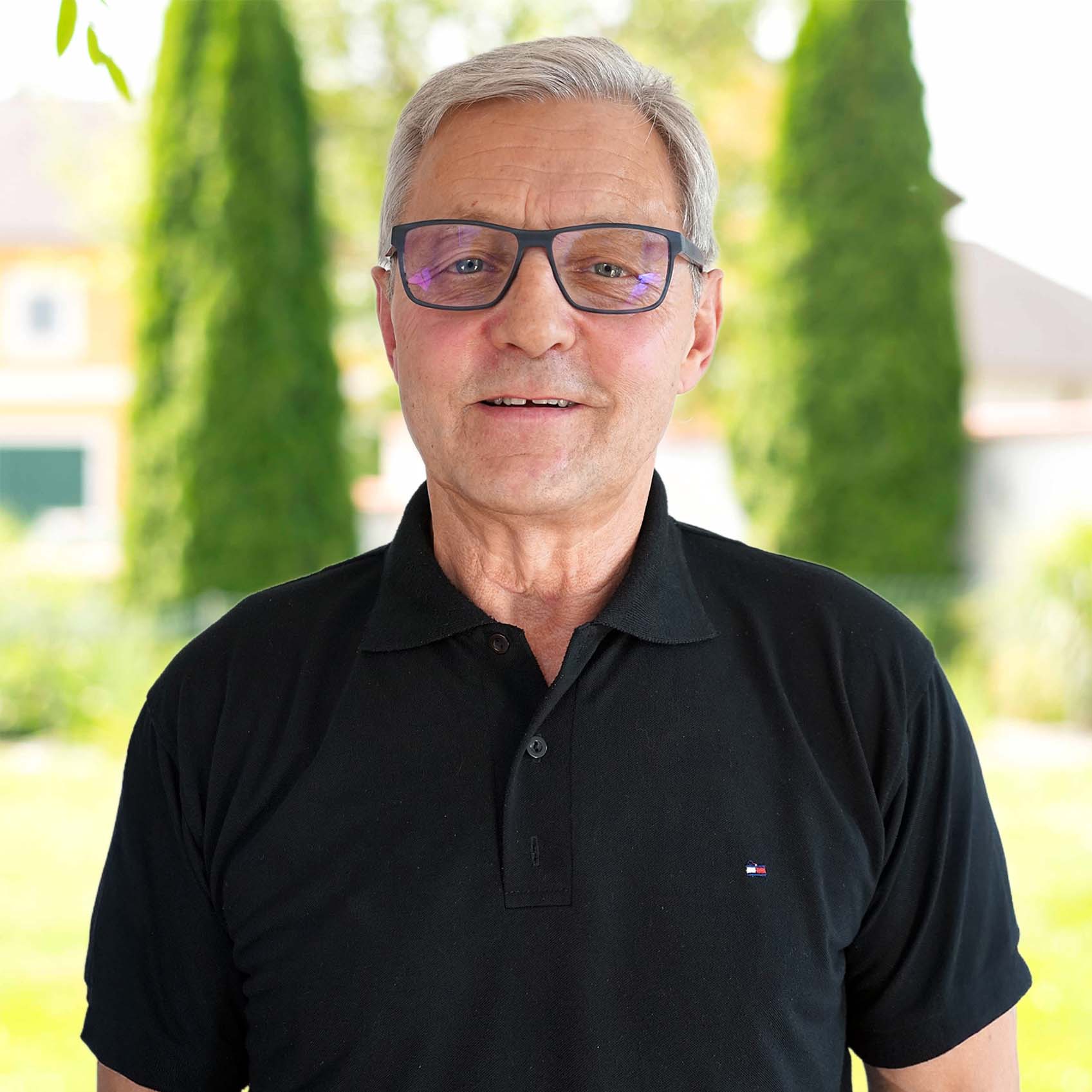 Älterer Mann mit grauen Haaren und Brille trägt ein schwarzes Polohemd vor einem verschwommenen Gartenhintergrund.