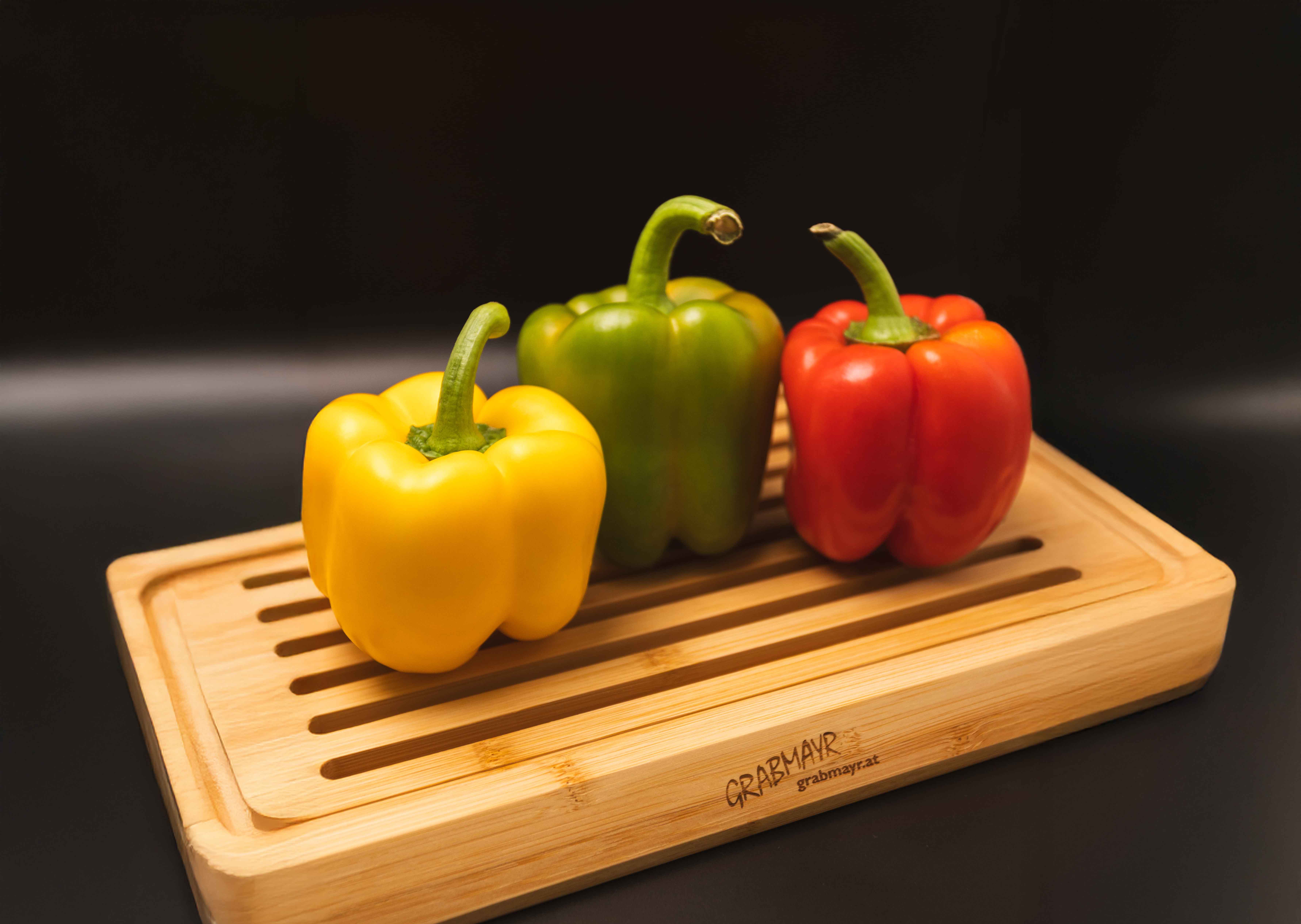 Drei Paprika in den Farben Gelb, Grün und Rot, die auf einem Holzbrett mit Schlitzablauf stehen.
