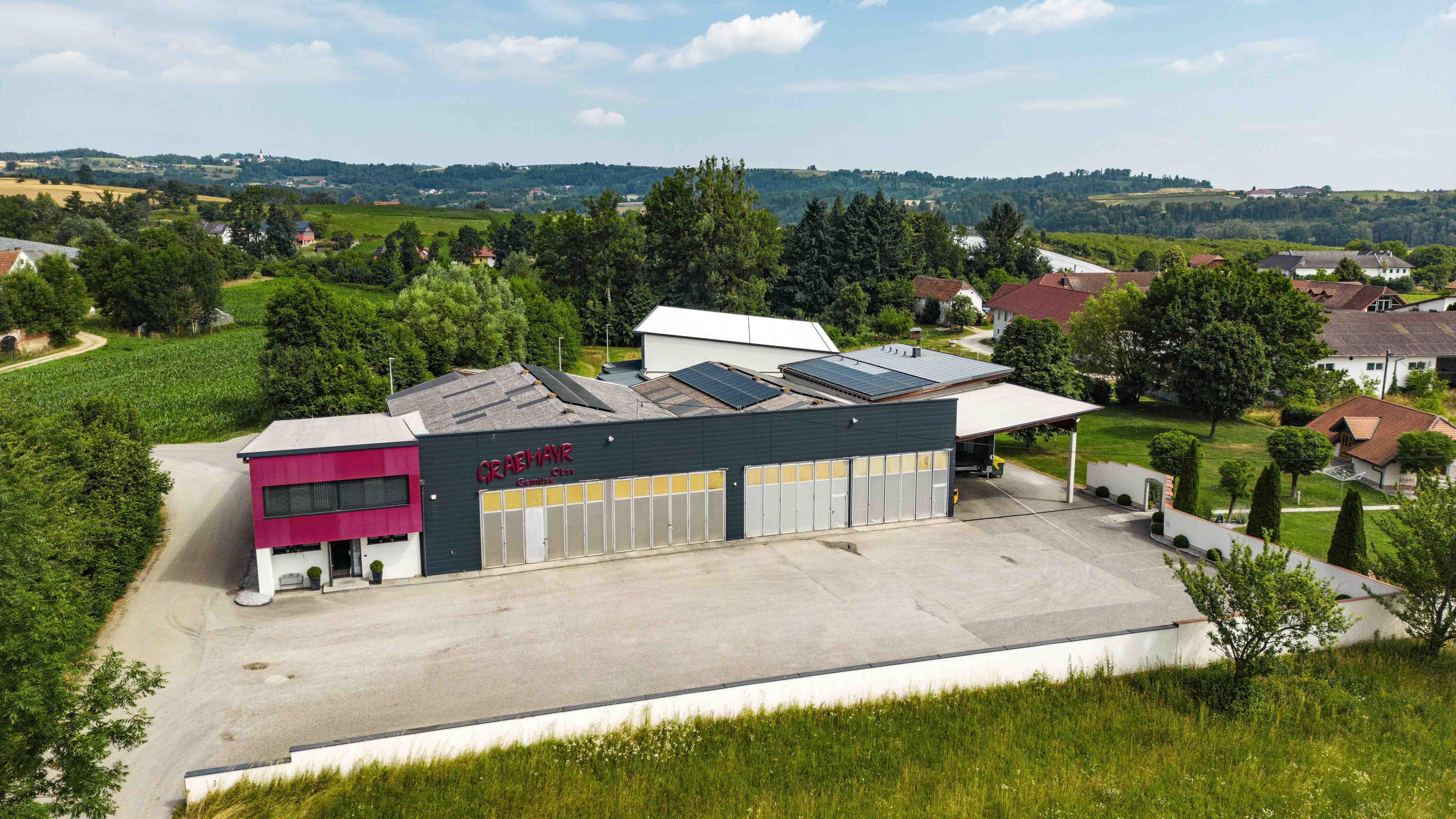 Industriegebäude mit Solaranlagen auf dem Dach, umgeben von grünen Feldern und vereinzelten Wohnhäusern in ländlicher Umgebung.