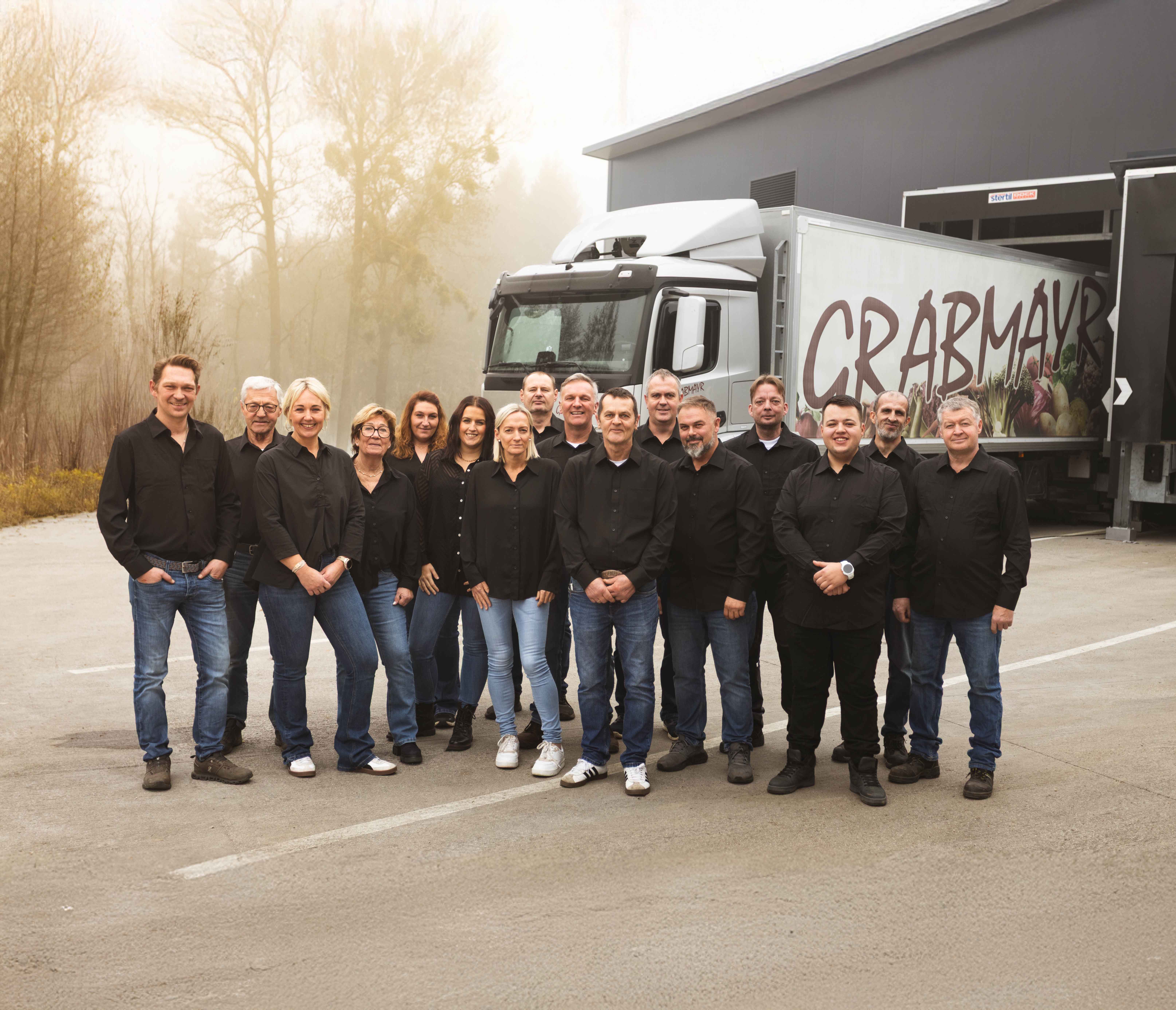 Gruppenfoto von 16 Personen in schwarzer Kleidung vor einem LKW mit der Aufschrift CRABMAYR bei einem Lagergebäude.