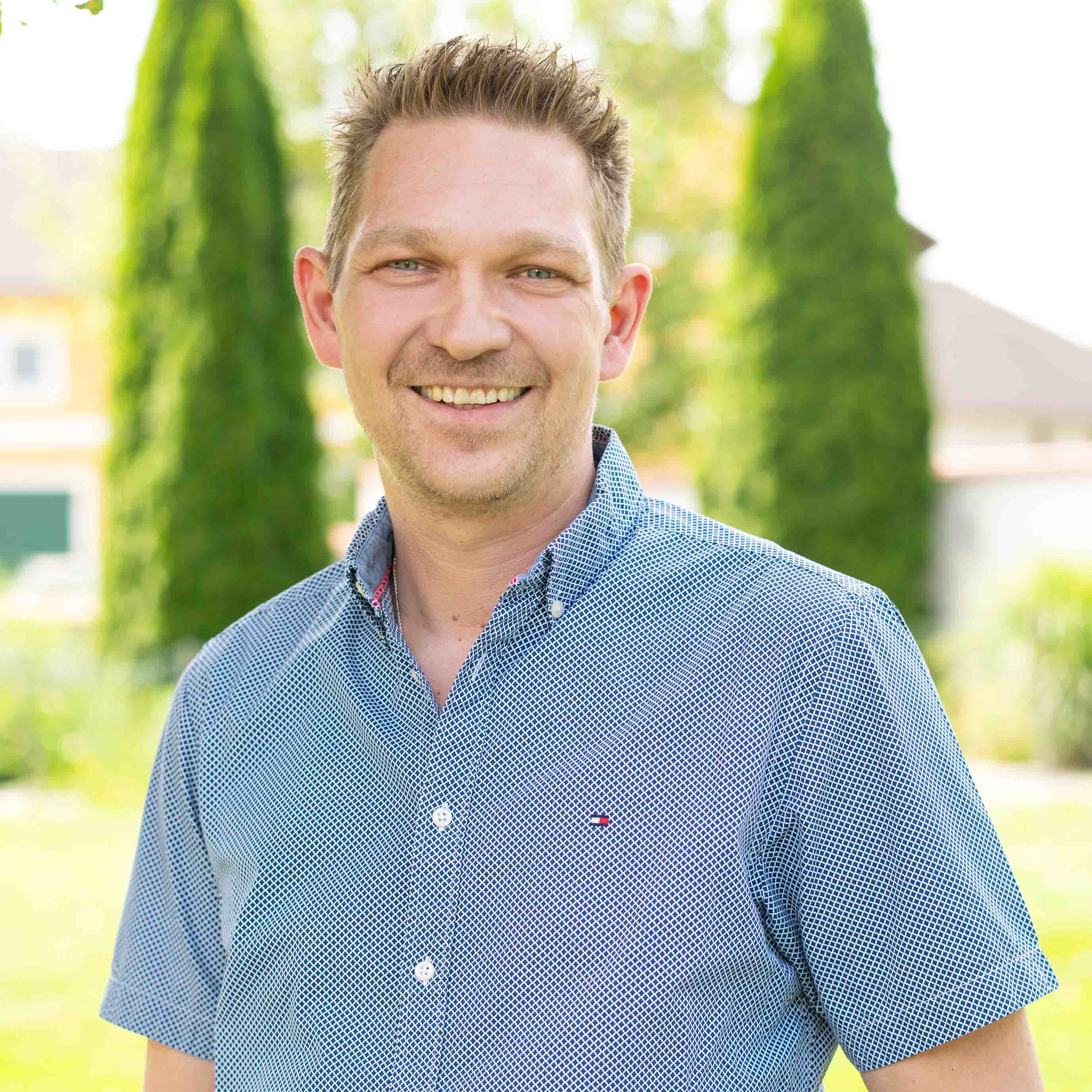 Lächelnder Mann mit kurz geschnittenem Haar trägt kurzärmliges, hellblau kariertes Hemd im Freien vor unscharfem Hintergrund mit Bäumen.