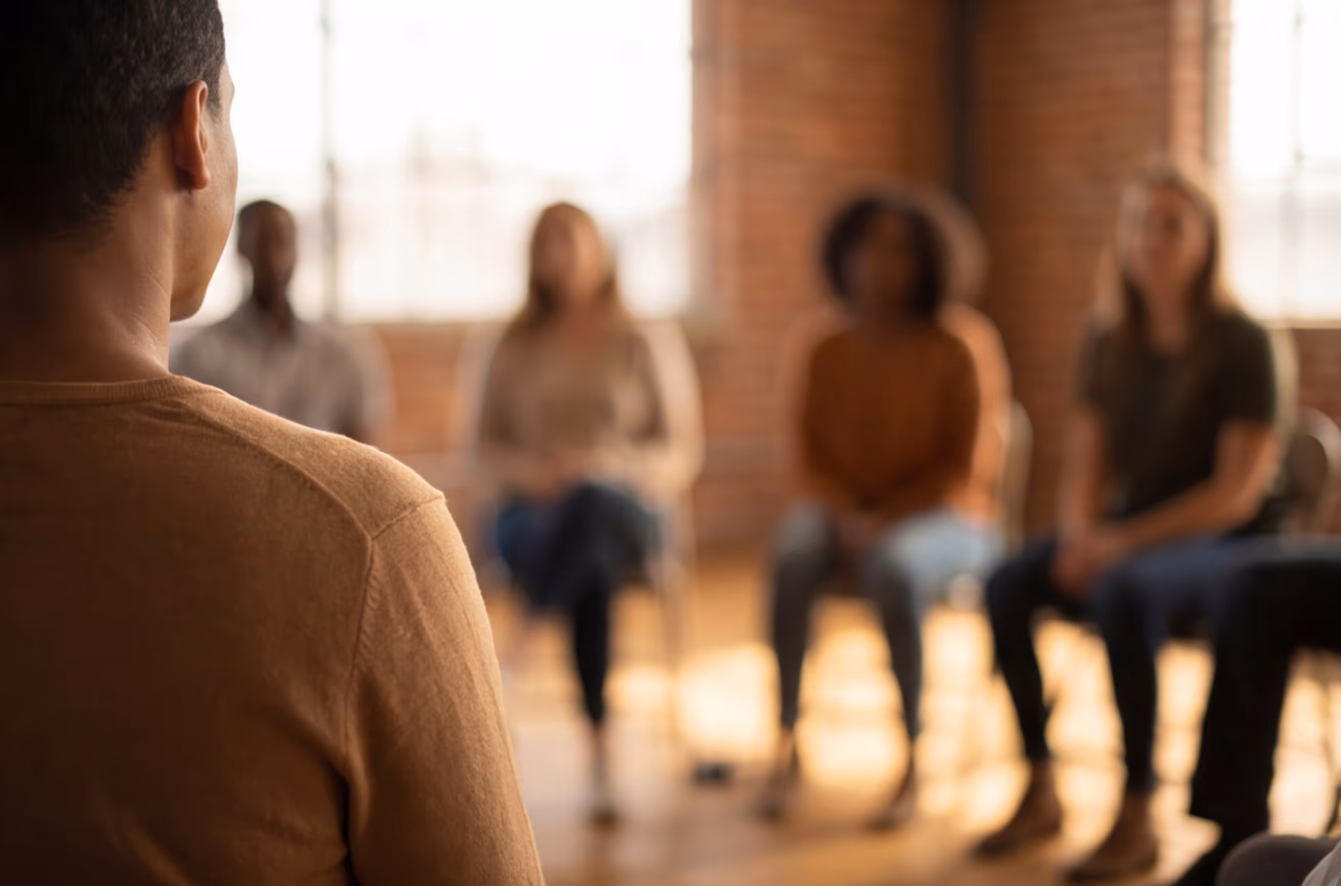 Group therapy session with a man speaking to four attentive people seated in a circle in a warmly lit room.