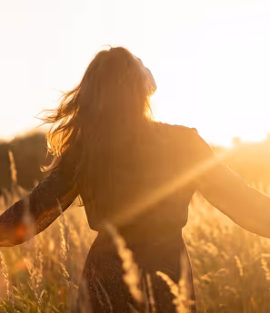 Person with long hair standing in a sunlit field with arms outstretched, backlit by golden sunlight.